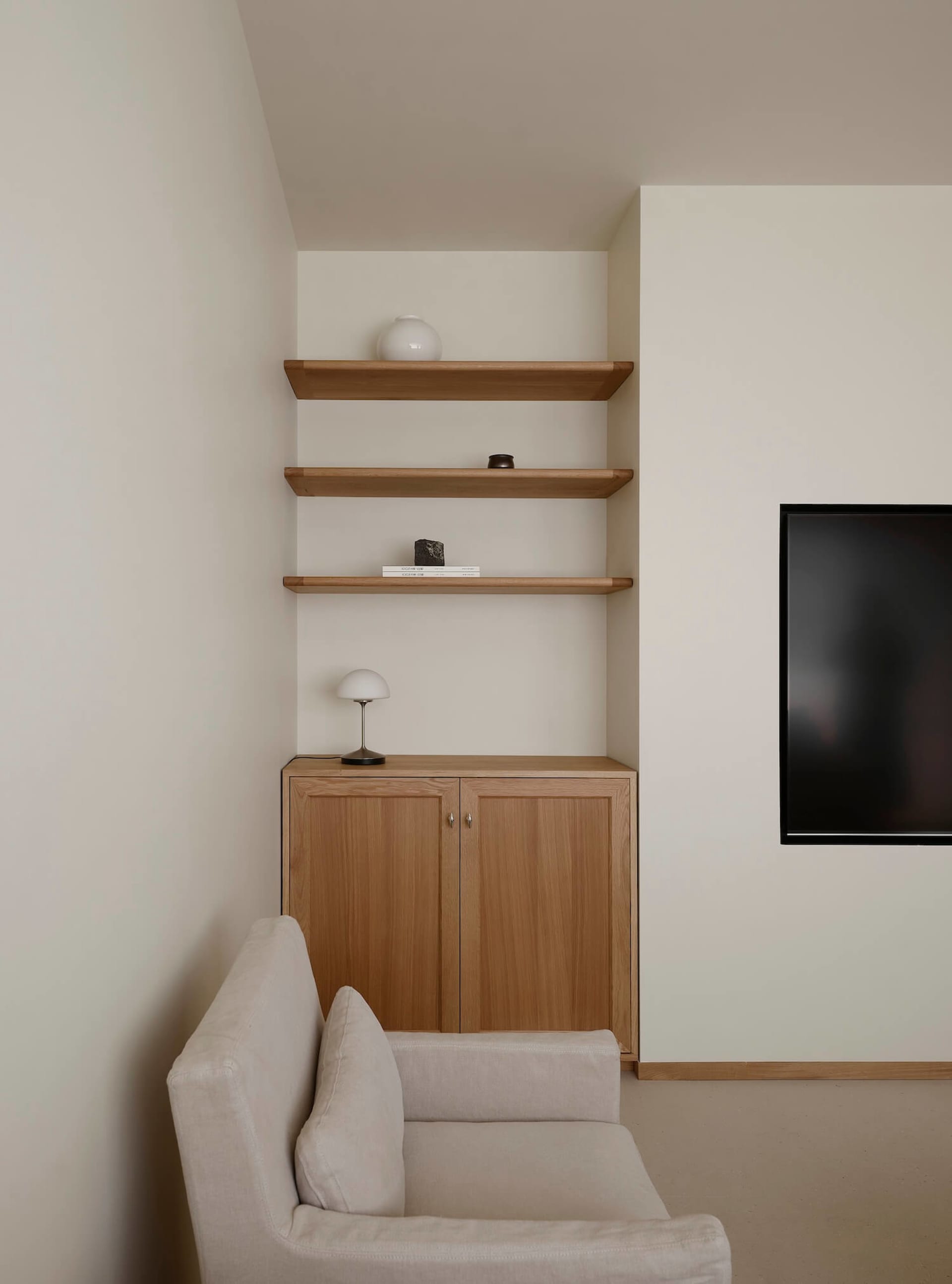 Minimalist living room corner with light oak cabinet, three floating oak shelves, white walls, and a cream fabric sofa in the foreground.