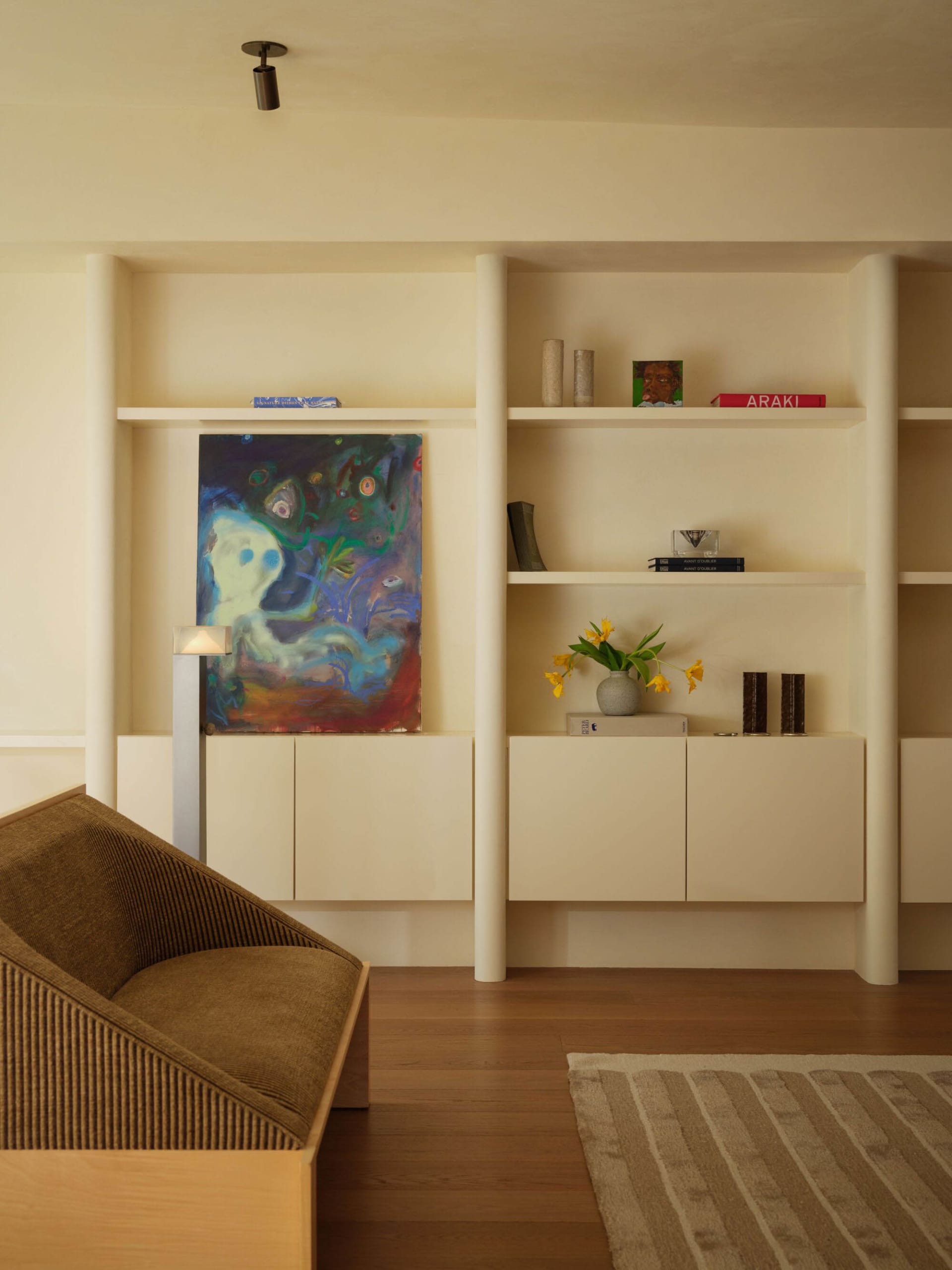 Modern minimalist living room with light oak flooring, built-in white shelving, a brown ribbed fabric armchair, colorful abstract painting, and decorative objects.