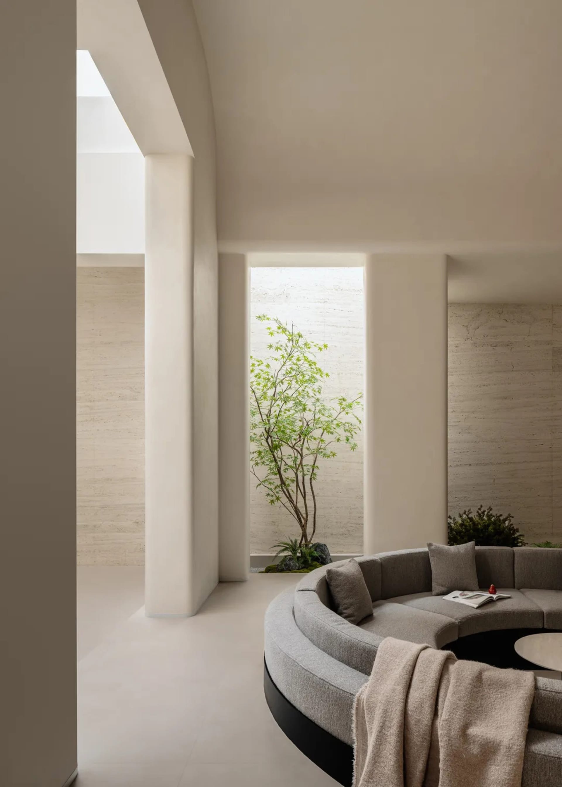 Modern minimalist living room featuring a large curved grey sectional sofa, travertine stone walls and floor, and an indoor courtyard with a Japanese maple tree.