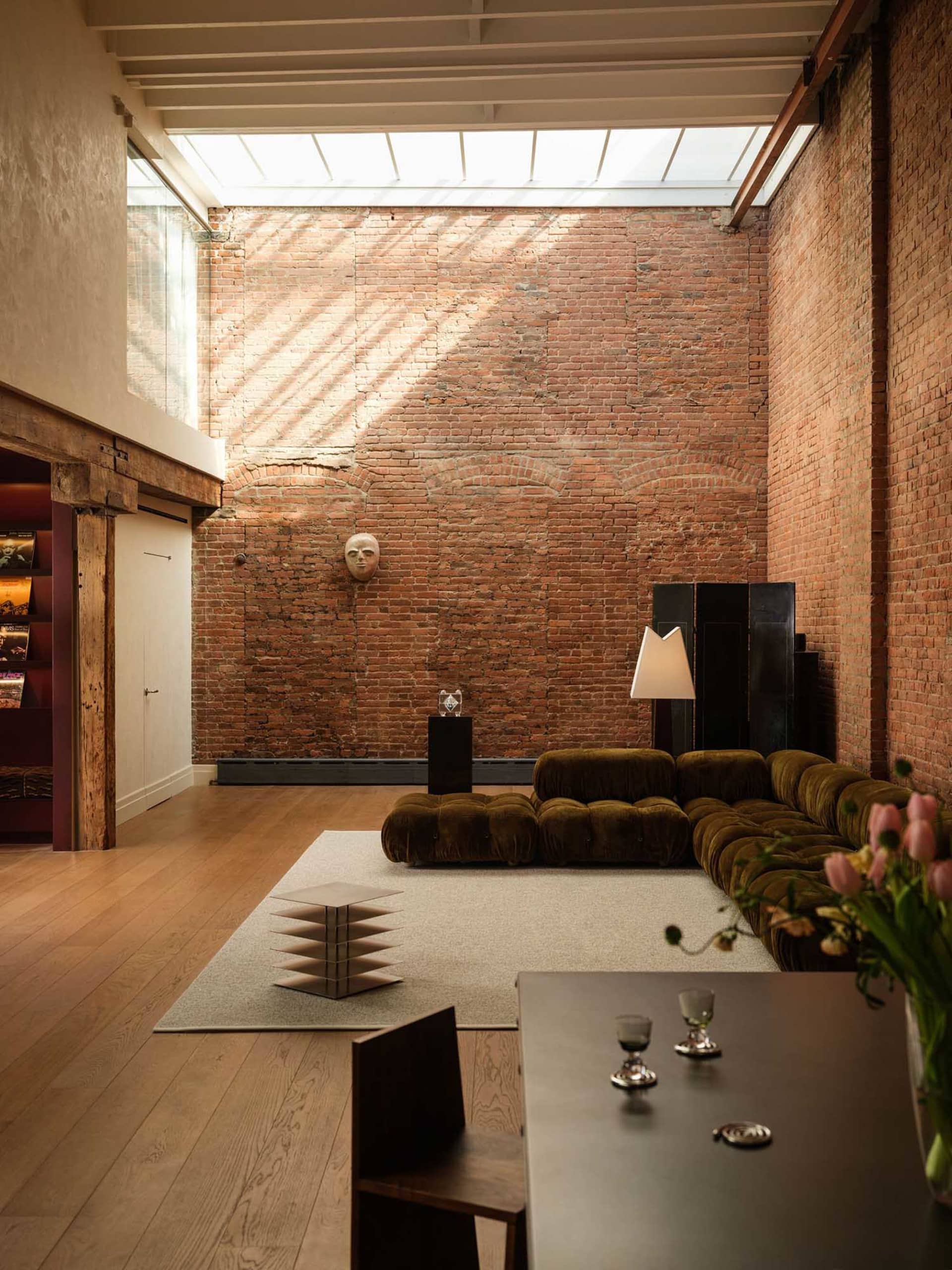 Spacious industrial loft living room featuring a full-height exposed red brick wall, olive green modular velvet sofa, light wood flooring, and a large skylight overhead.
