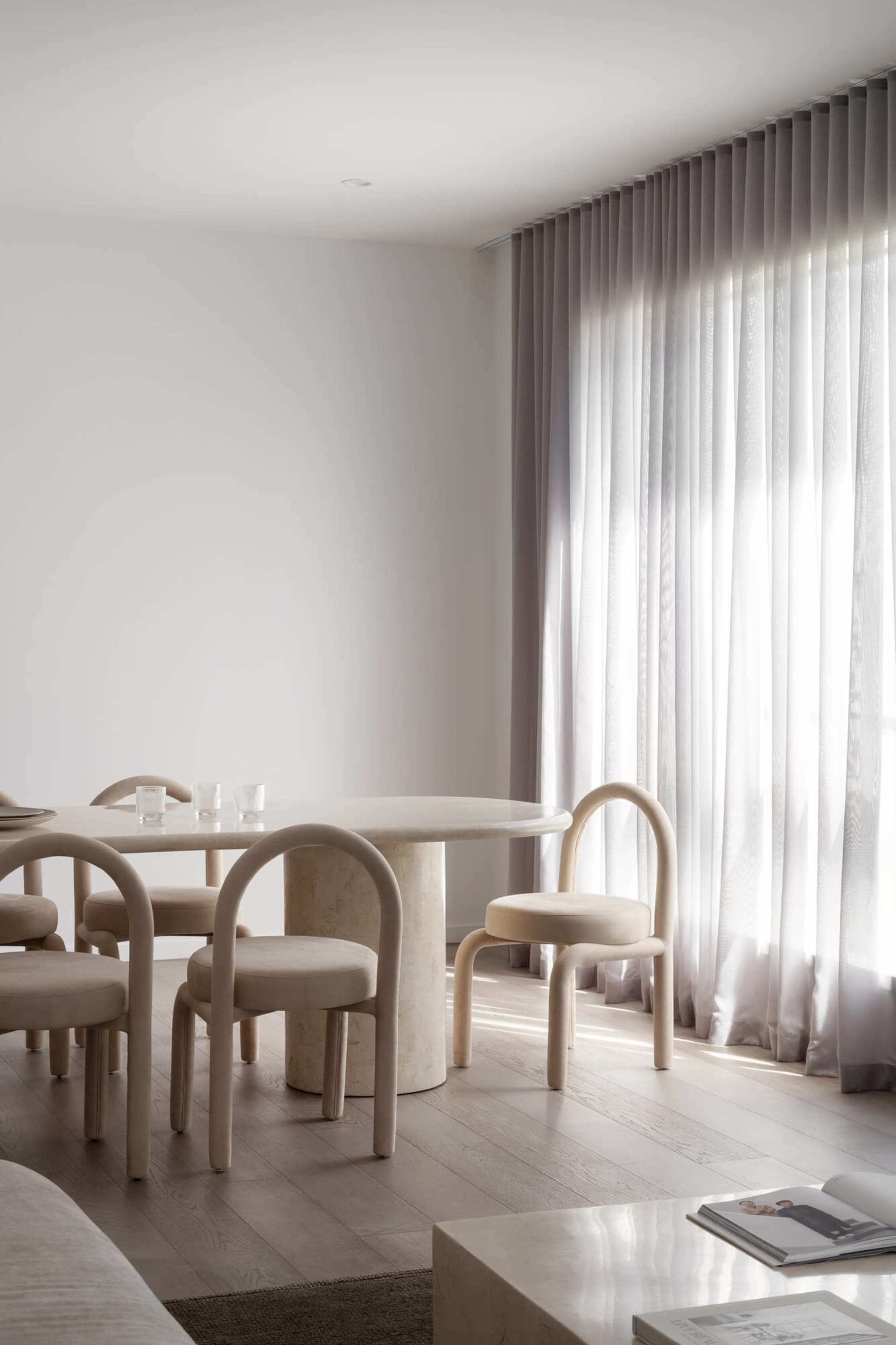 Minimalist dining room featuring a round travertine table, curved beige upholstered chairs, light wood flooring, and floor-to-ceiling sheer curtains with bright natural light.