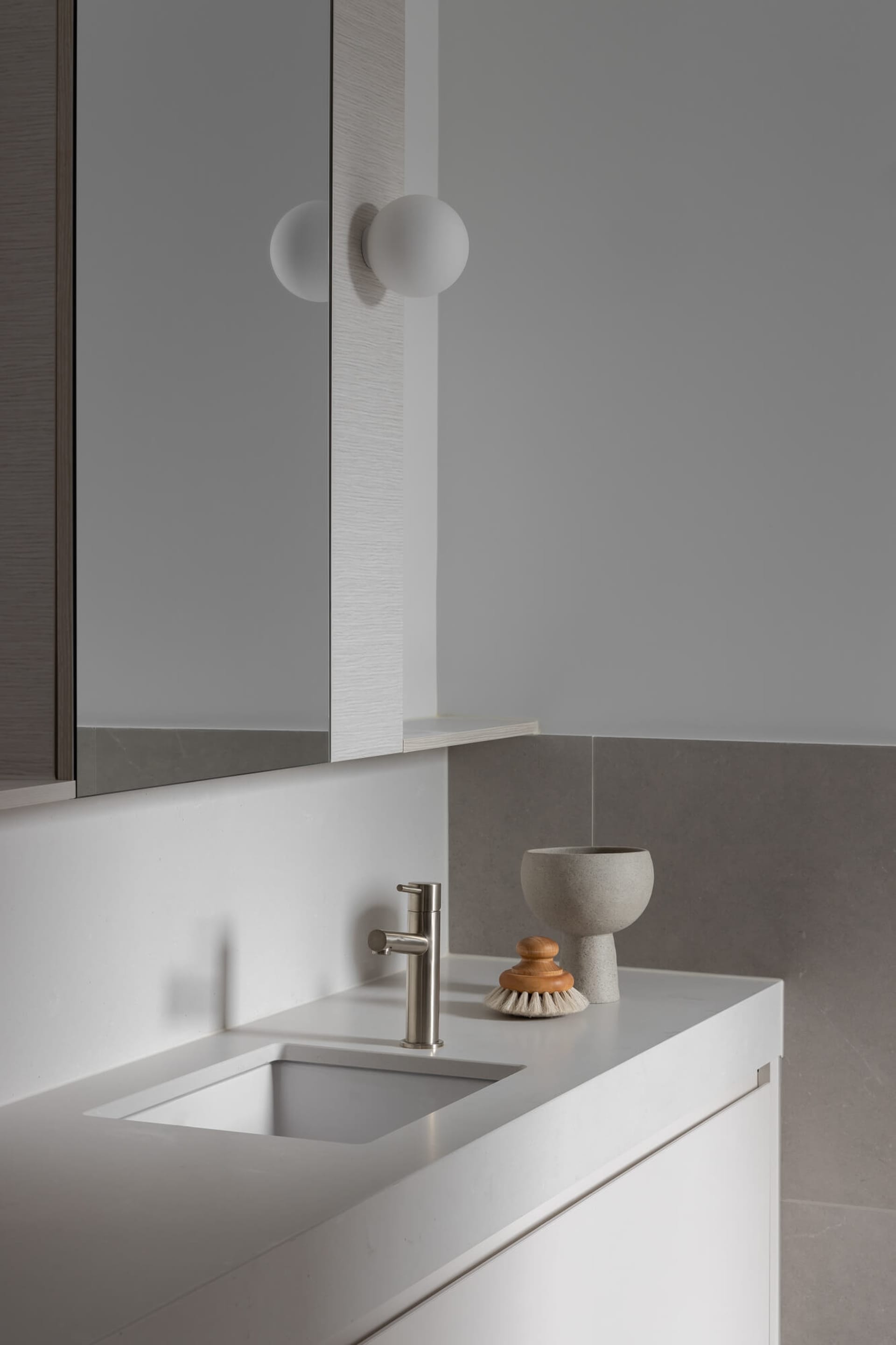 Modern minimalist bathroom vanity featuring a white integrated sink, brushed nickel faucet, concrete bowl, wooden brush, and two spherical wall lamps against grey tiled walls.