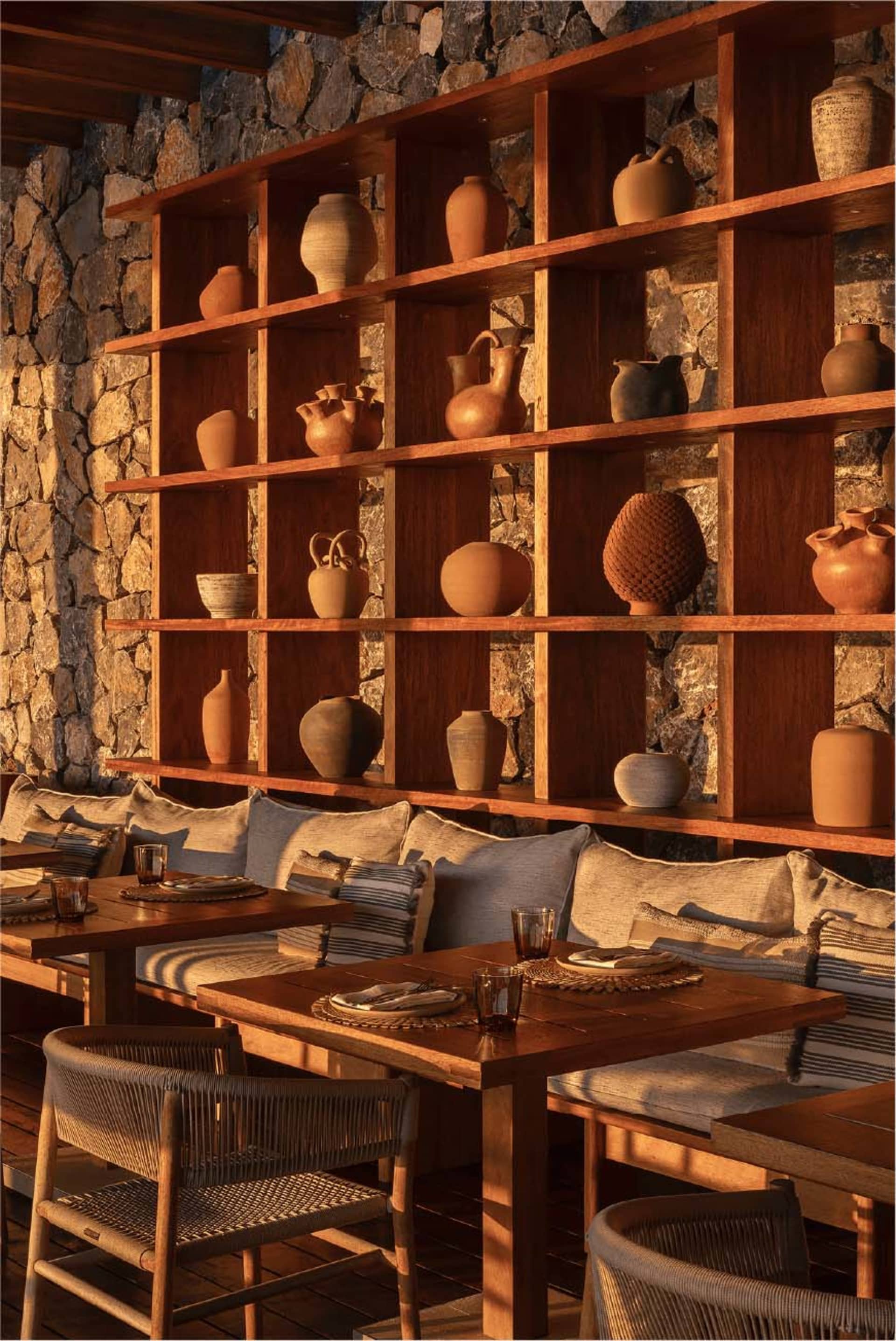 Cozy rustic dining area featuring a built-in wooden shelving unit filled with terracotta pottery, set against a natural stone wall with cushioned banquette seating and woven chairs.