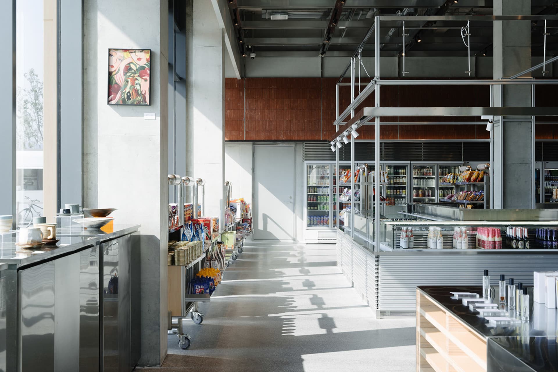 Modern industrial-style convenience store interior with polished concrete floors, stainless steel counters, open shelving, and a second-floor mezzanine structure.