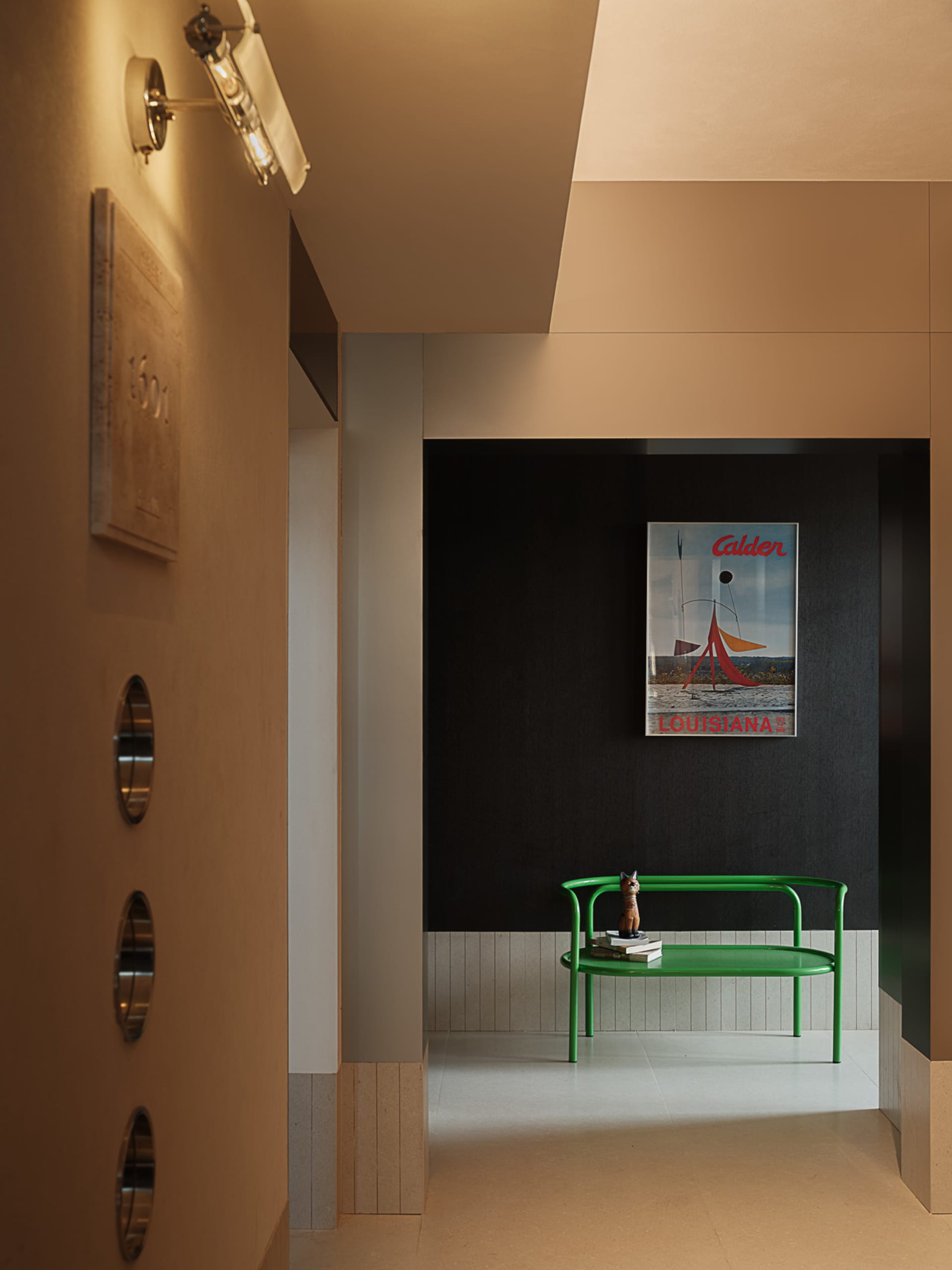 Modern minimalist entryway featuring a bright green metal bench, Alexander Calder Louisiana poster, warm wall sconce, and two-tone beige and dark grey walls with wood accents.