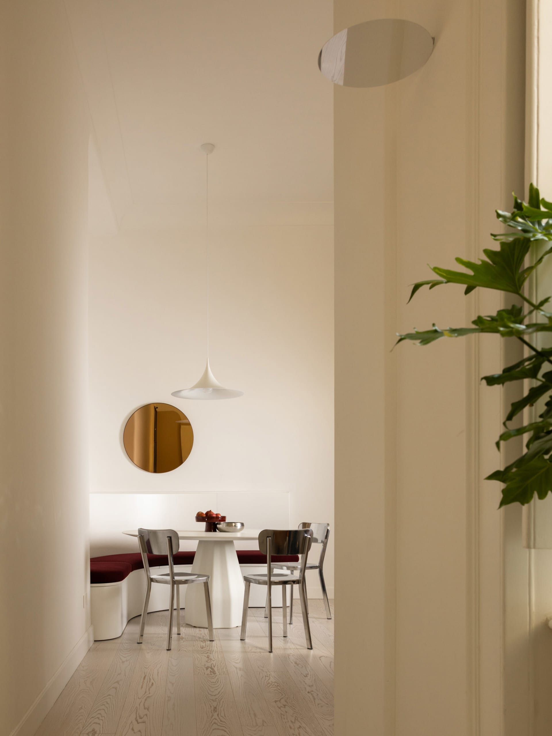 Modern minimalist dining area featuring a curved white banquette with red velvet upholstery, a white pedestal table, transparent acrylic and stainless steel chairs, a white pendant lamp, and a round brass mirror on the wall.