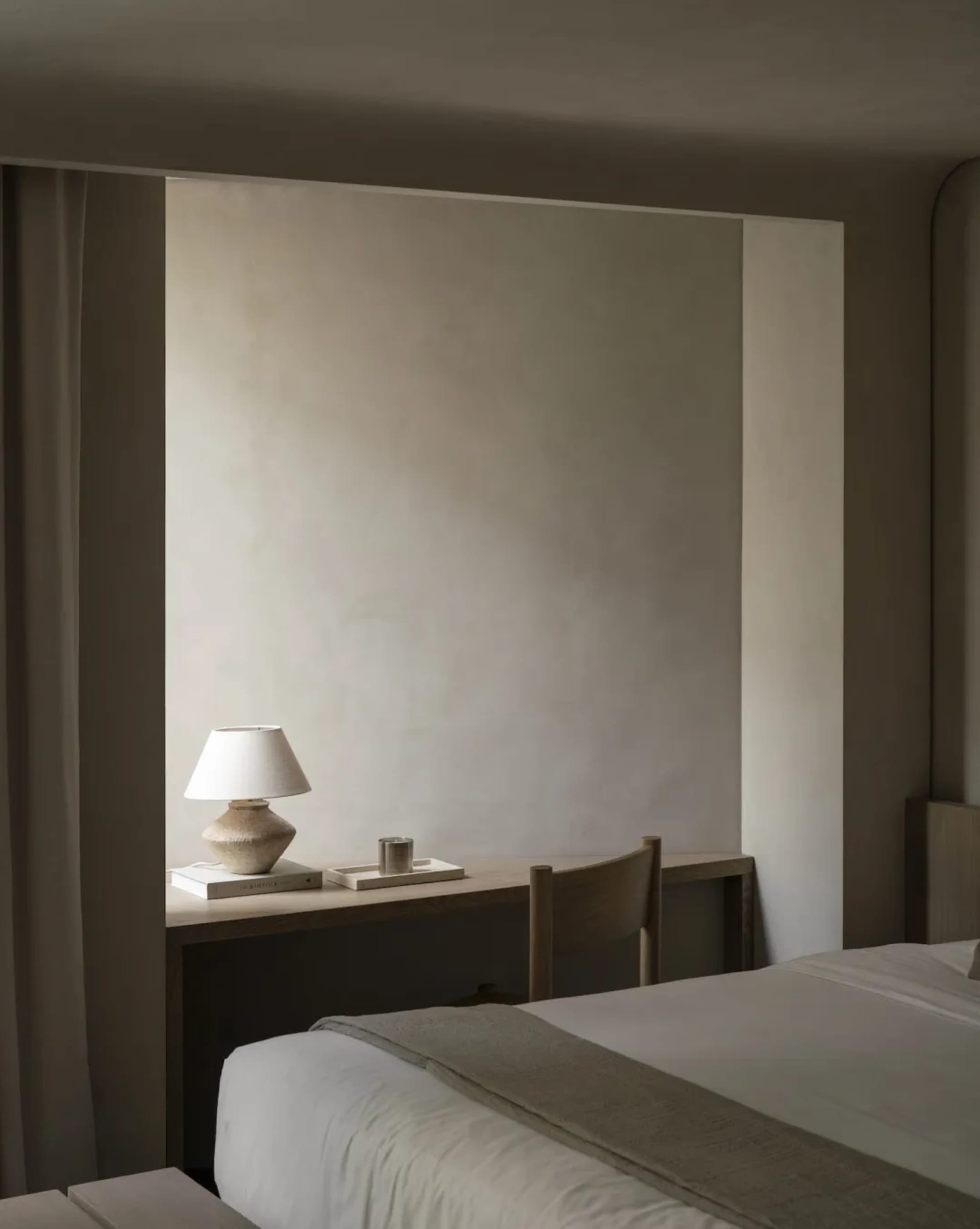 Minimalist bedroom featuring a built-in oak desk with a ceramic table lamp, wooden chair, and neutral bedding in soft beige tones.