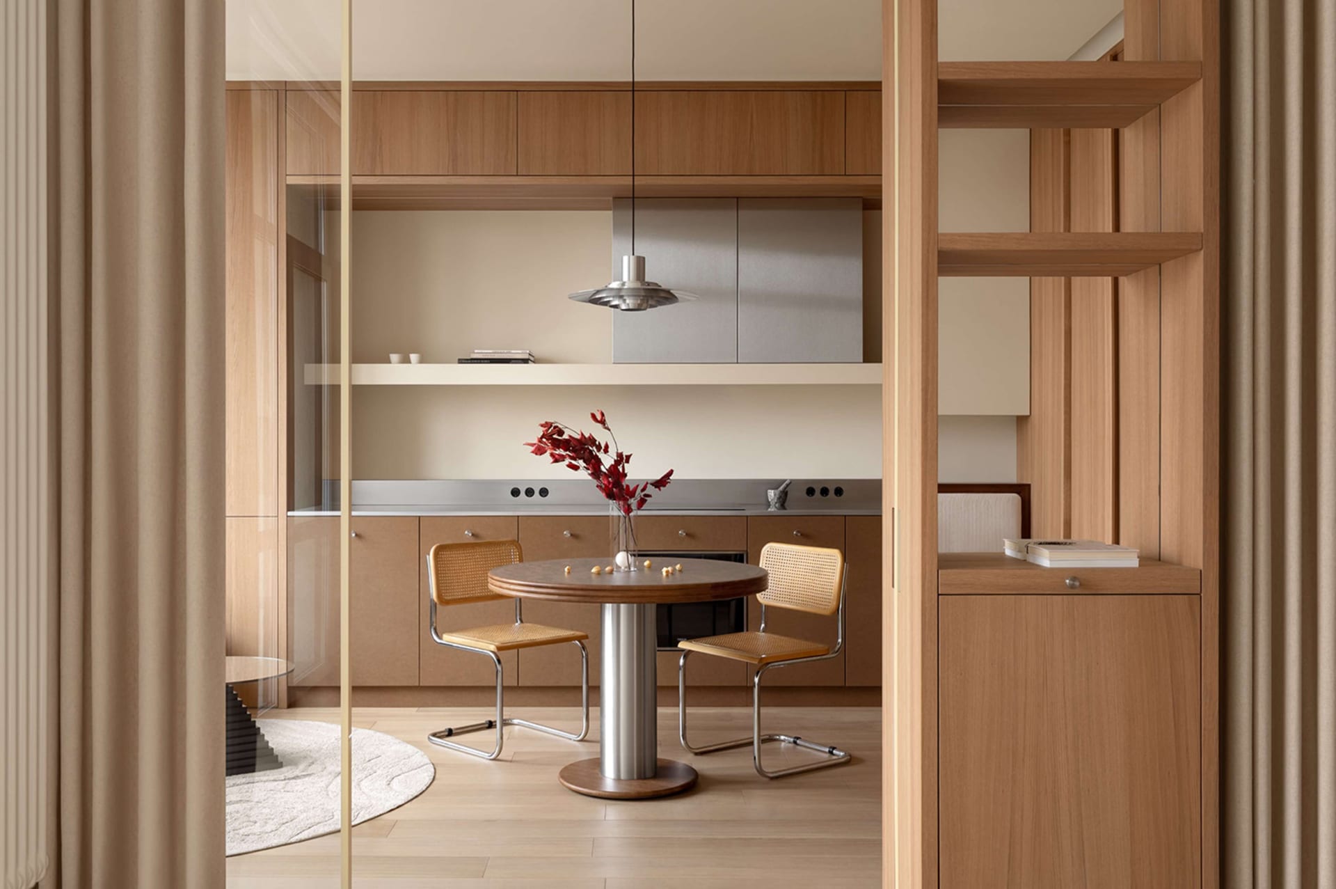 Modern minimalist dining area featuring a round wooden table, two cane and metal chairs, warm oak cabinetry, and a stainless steel pendant light.
