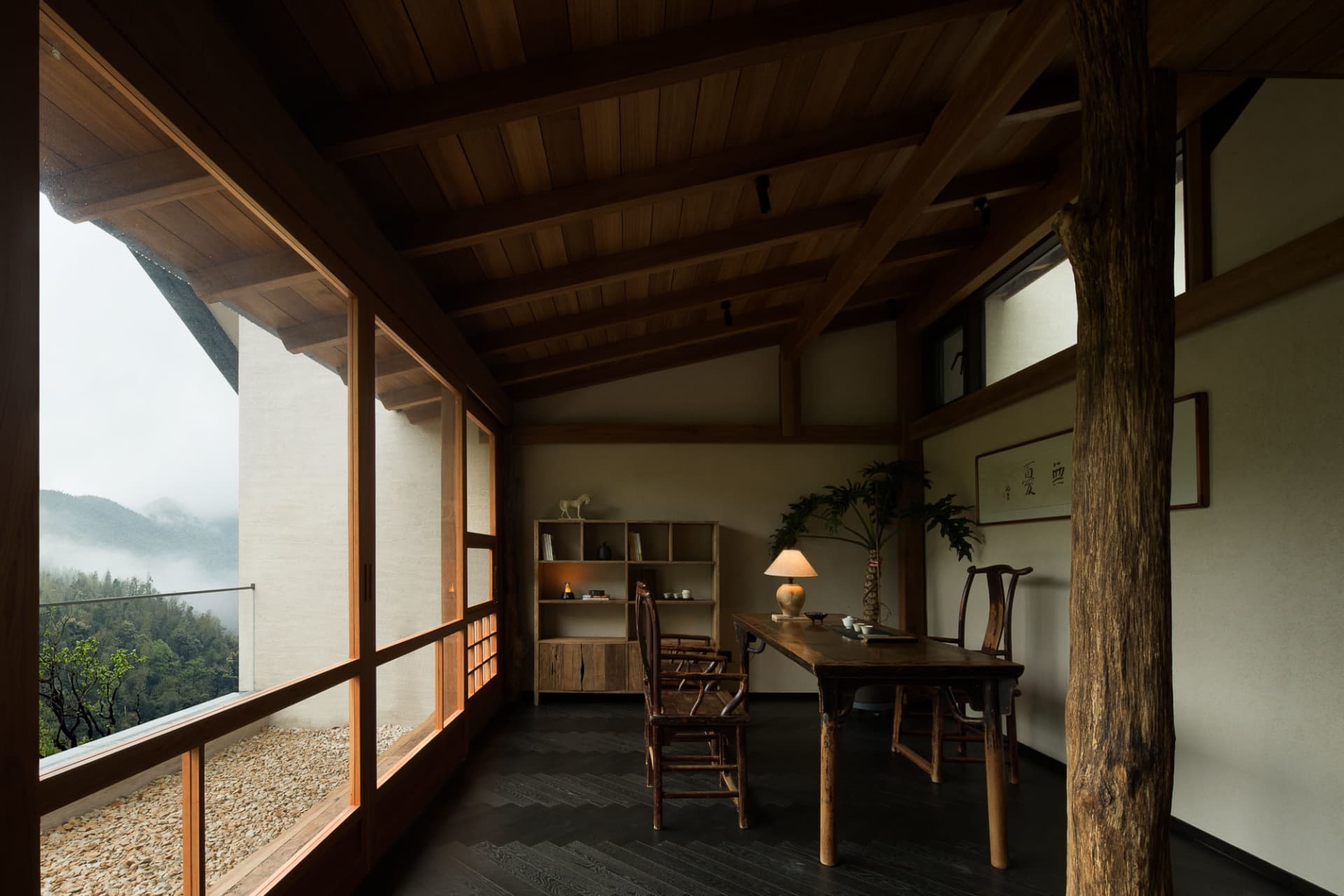 Interior of a mountain villa dining room featuring solid wood furniture, exposed timber ceiling beams, and large windows revealing a misty forest landscape.