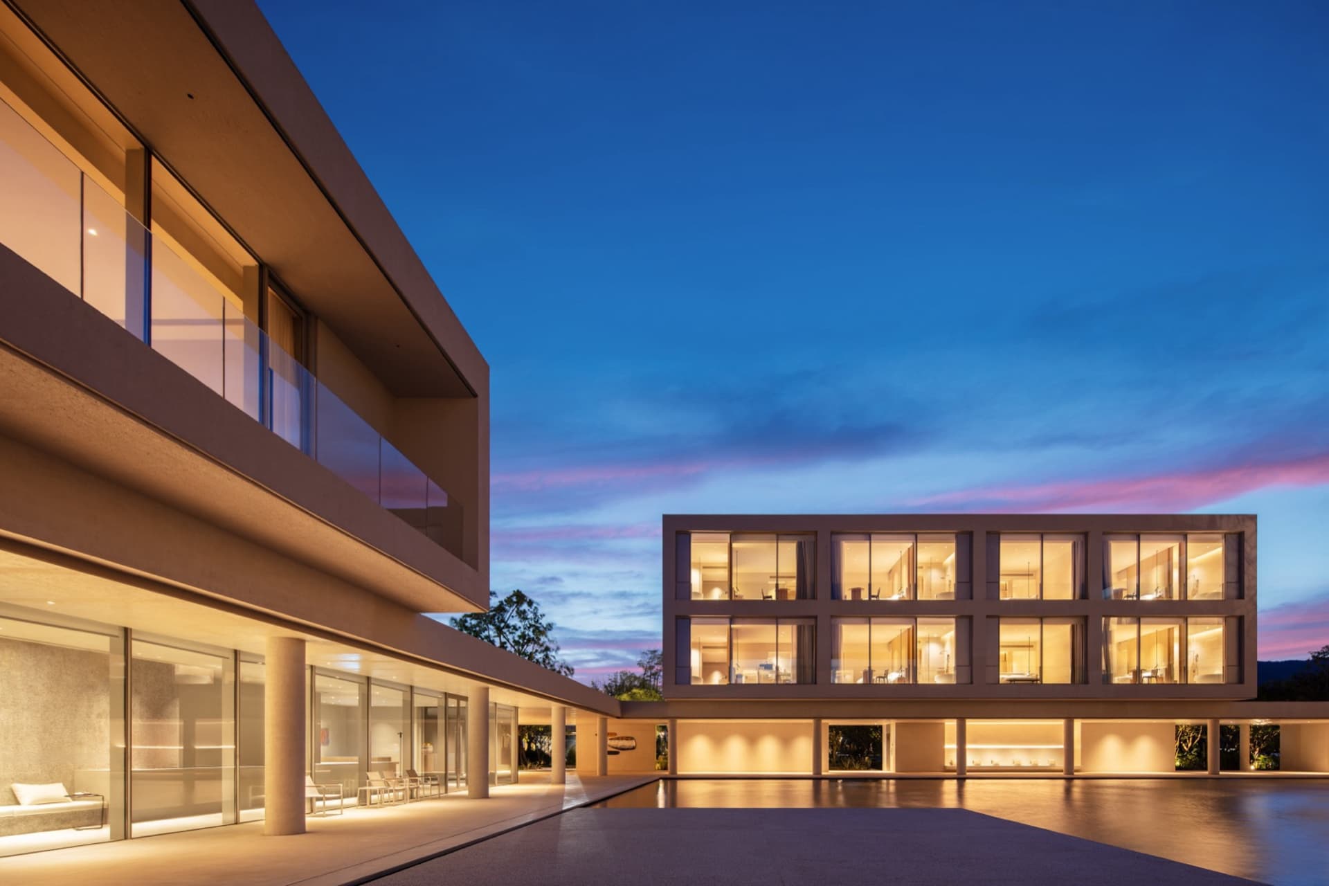 Modern two-story concrete house with extensive glass windows glowing warmly at twilight, featuring a large reflecting pool in the foreground and a clear blue-to-pink gradient sky.