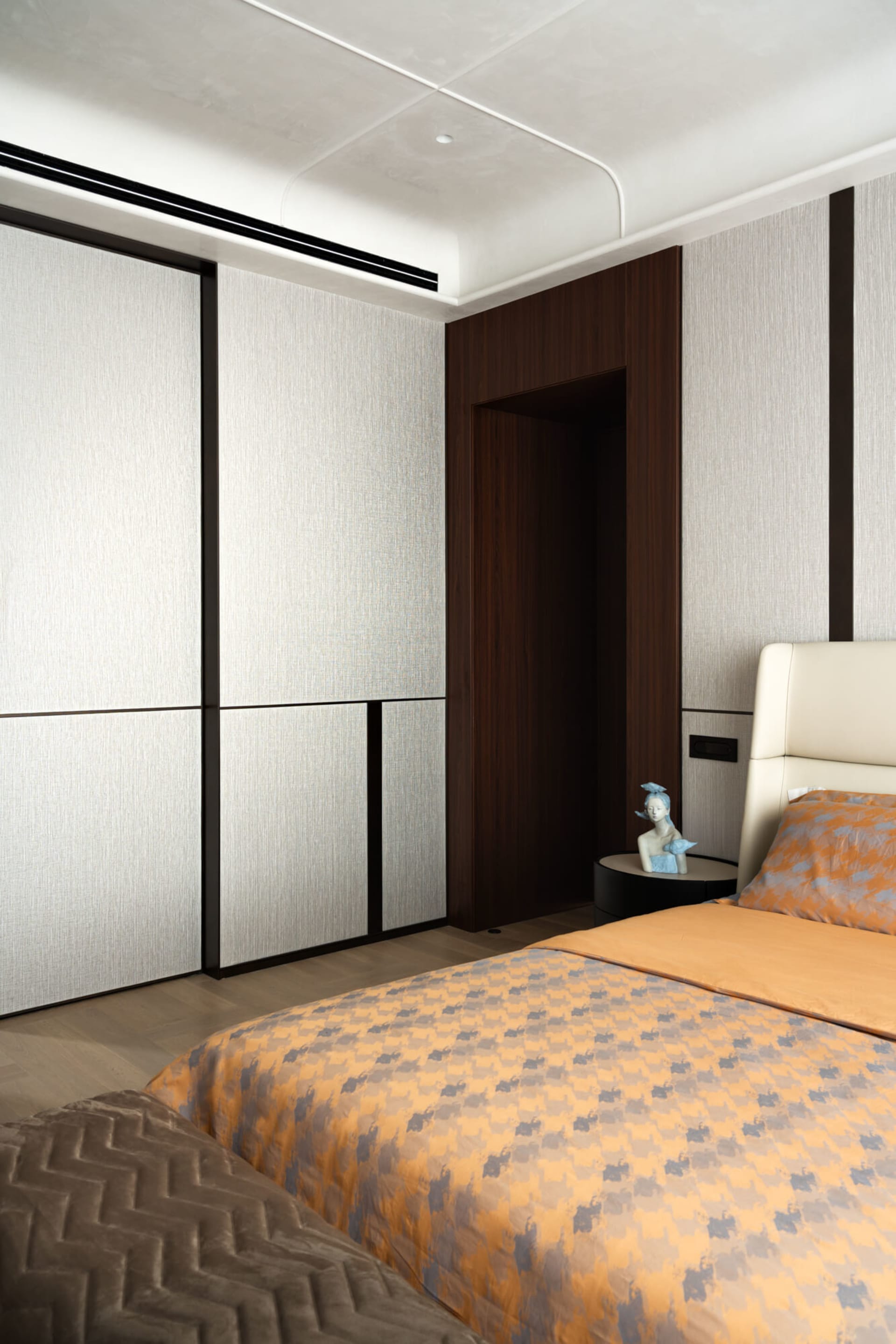 Contemporary bedroom interior with dark walnut built-in wardrobe, textured light grey walls, cream leather headboard, and orange and grey geometric patterned bedding on a low platform bed.