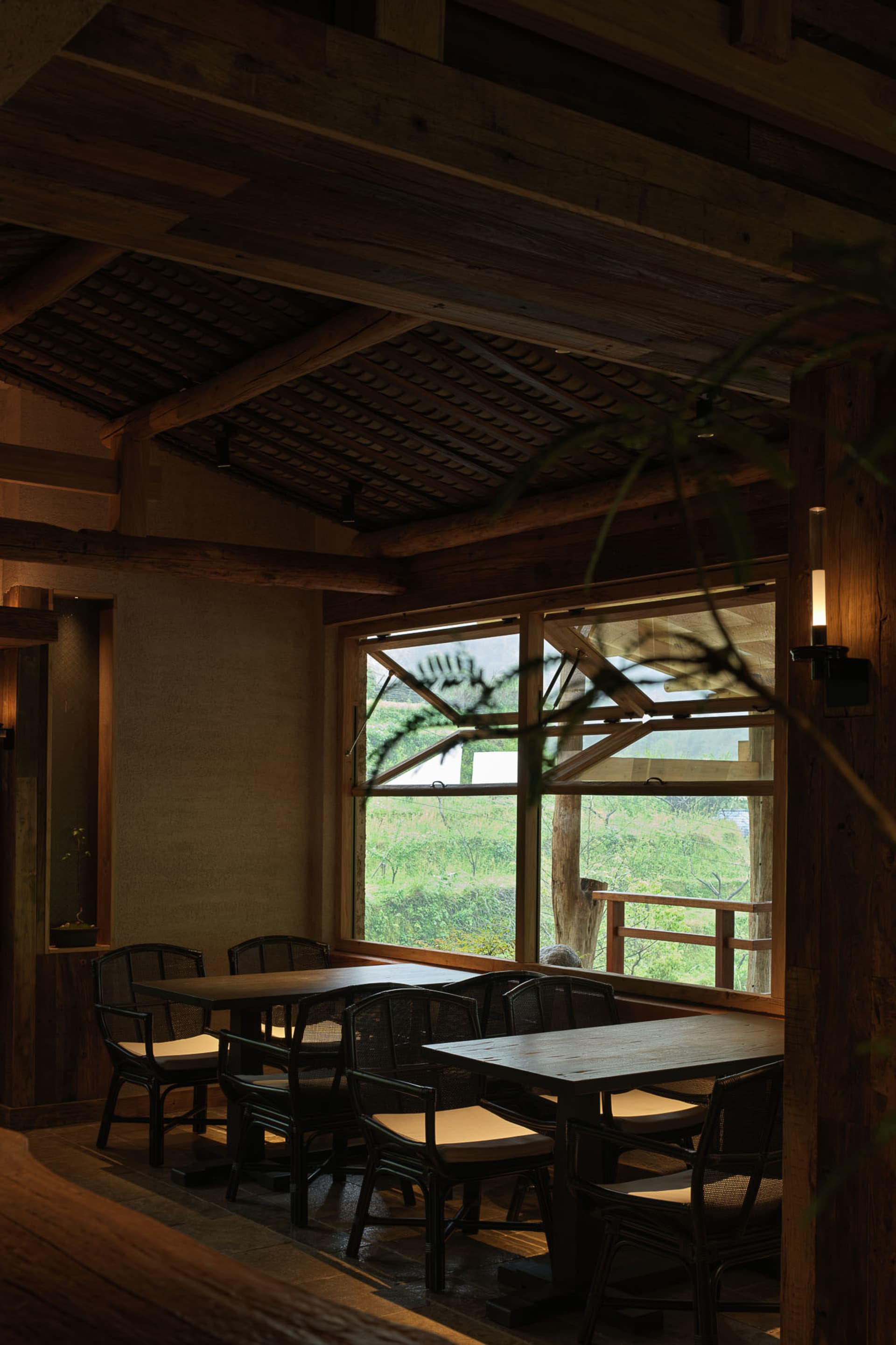 Interior of a rustic dining room featuring reclaimed wood beams, dark wooden tables, rattan dining chairs, and large windows overlooking a lush green mountain landscape.