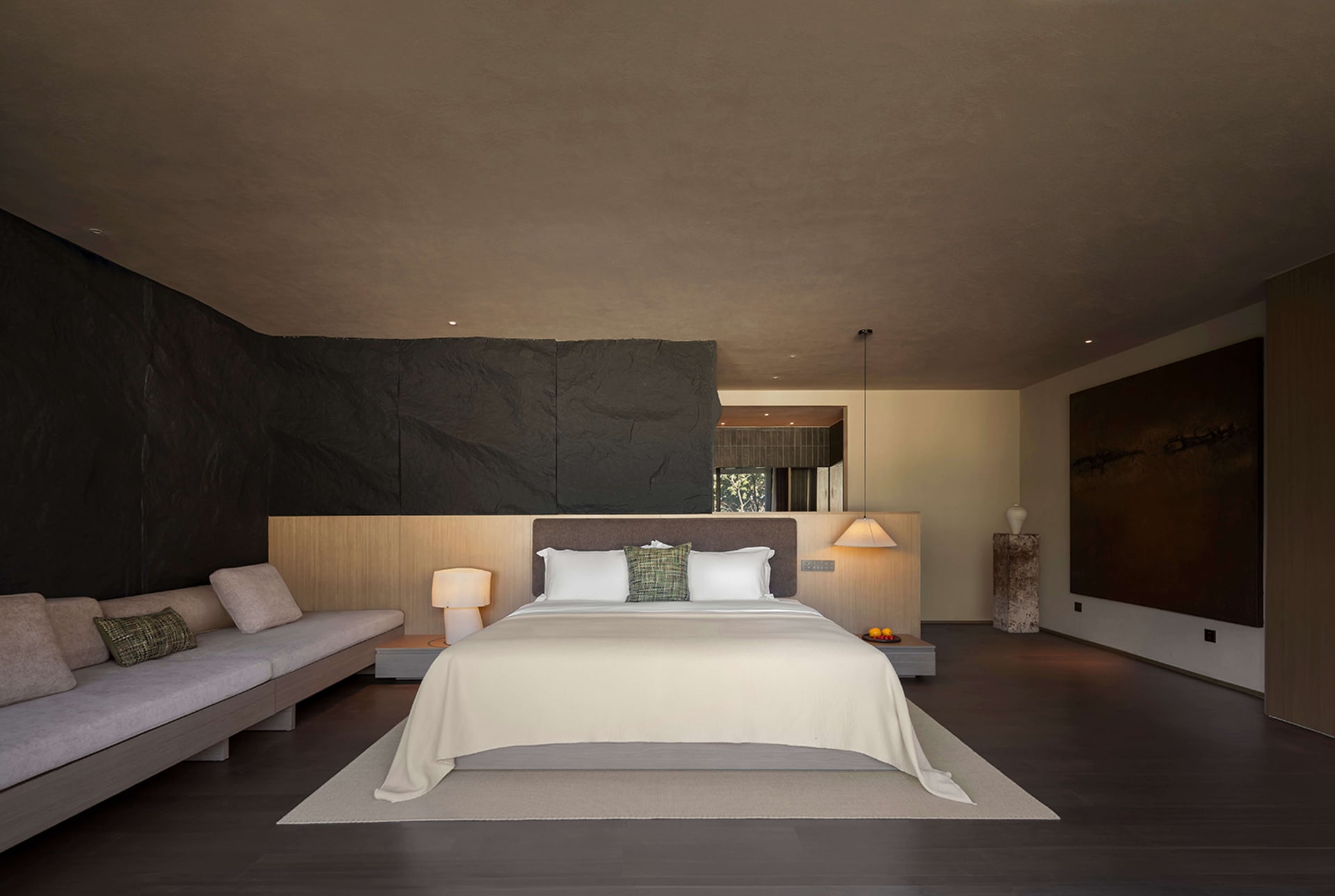 Modern minimalist bedroom featuring a large dark slate stone feature wall, built-in daybed with neutral cushions, king size bed with white linens, pendant light, and large abstract painting.