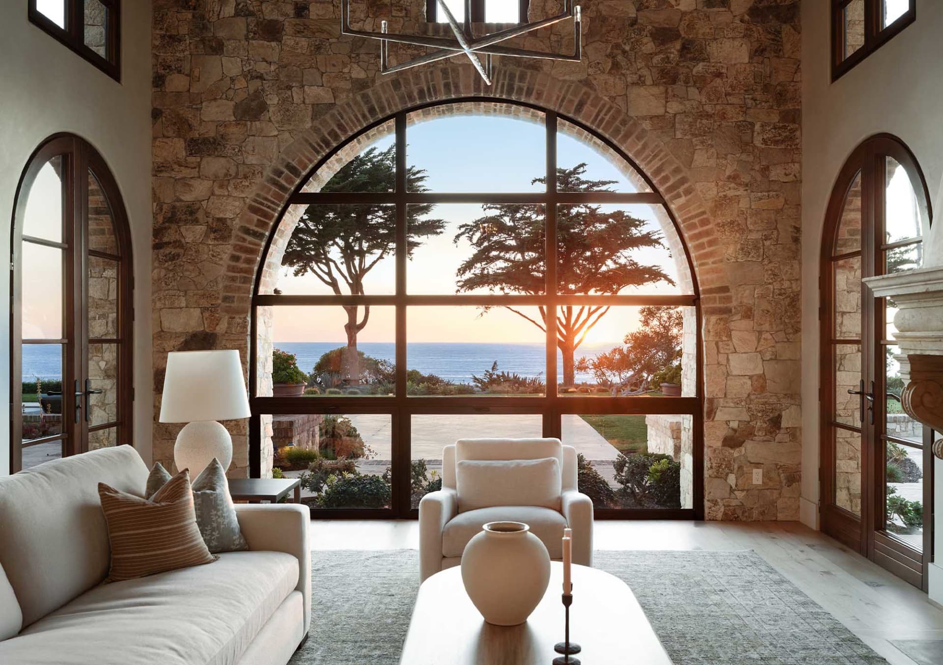 Luxurious living room featuring a massive arched picture window with ocean view at sunset, stone walls, cream sofa, and modern chandelier.