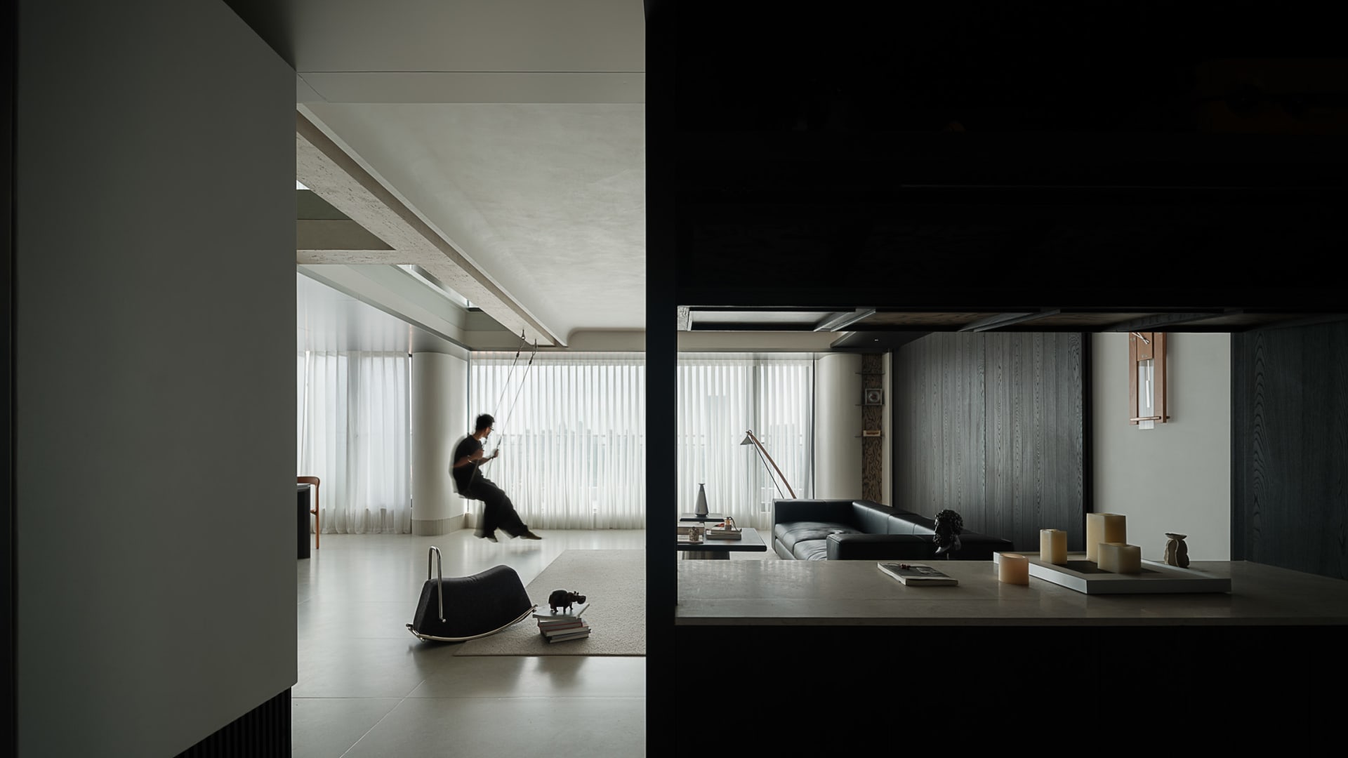 Modern minimalist living room featuring a hanging indoor swing, micro-cement walls and ceiling, dark wood paneling, and a black leather sofa under soft natural light.