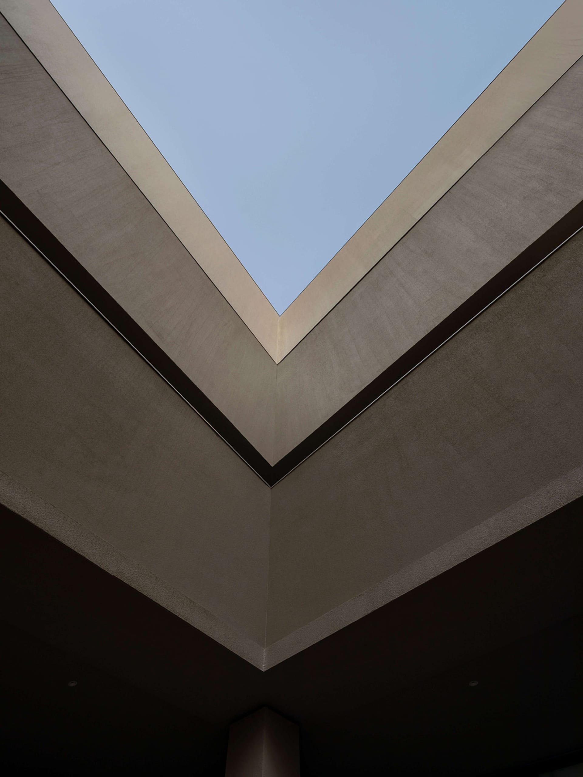 Looking straight up at a modern V-shaped concrete ceiling with a large rectangular skylight opening to blue sky.