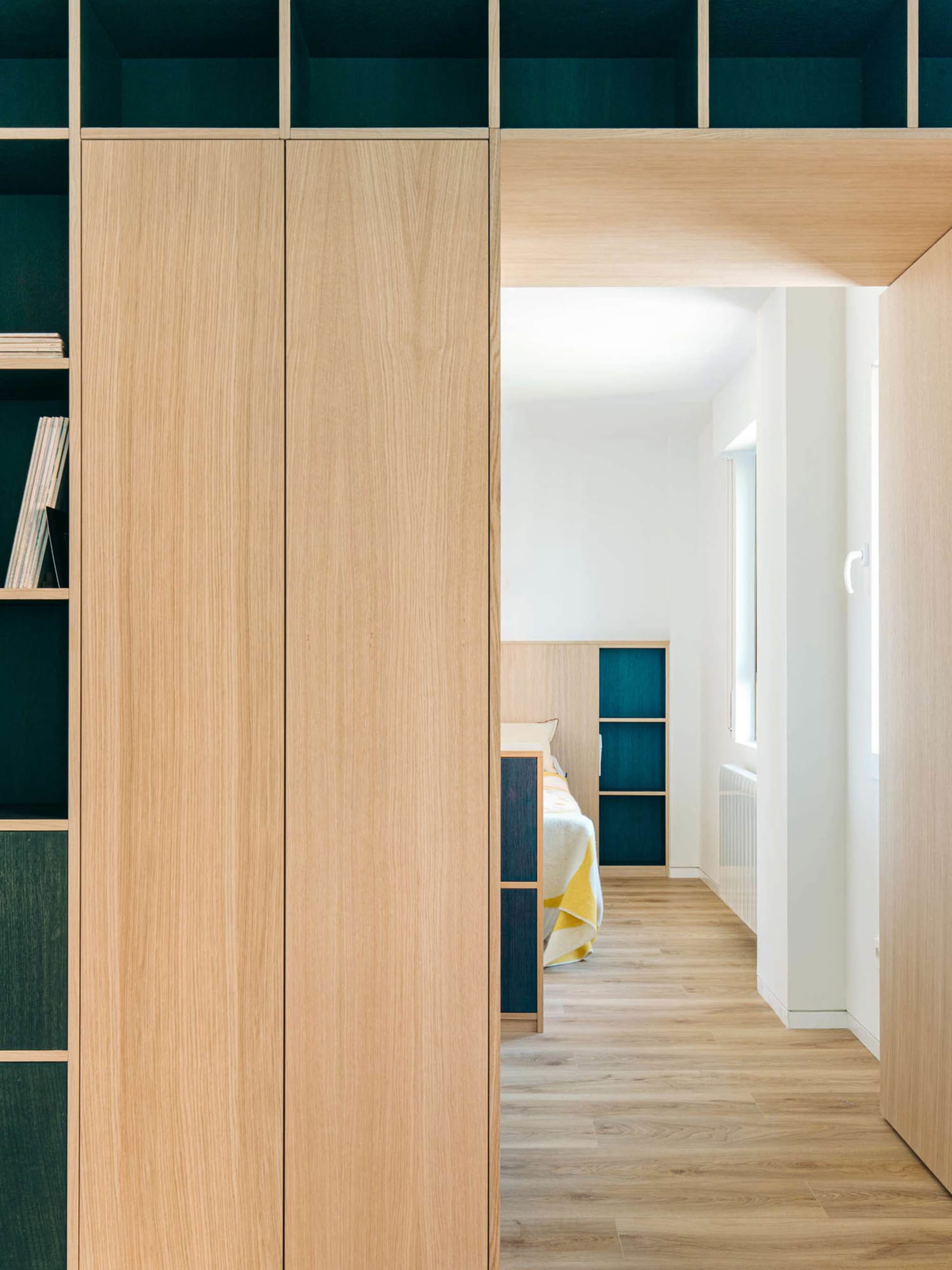 View from a hallway through a large oak built-in wardrobe into a modern bedroom with teal shelving niches and a bed with yellow throw blanket.