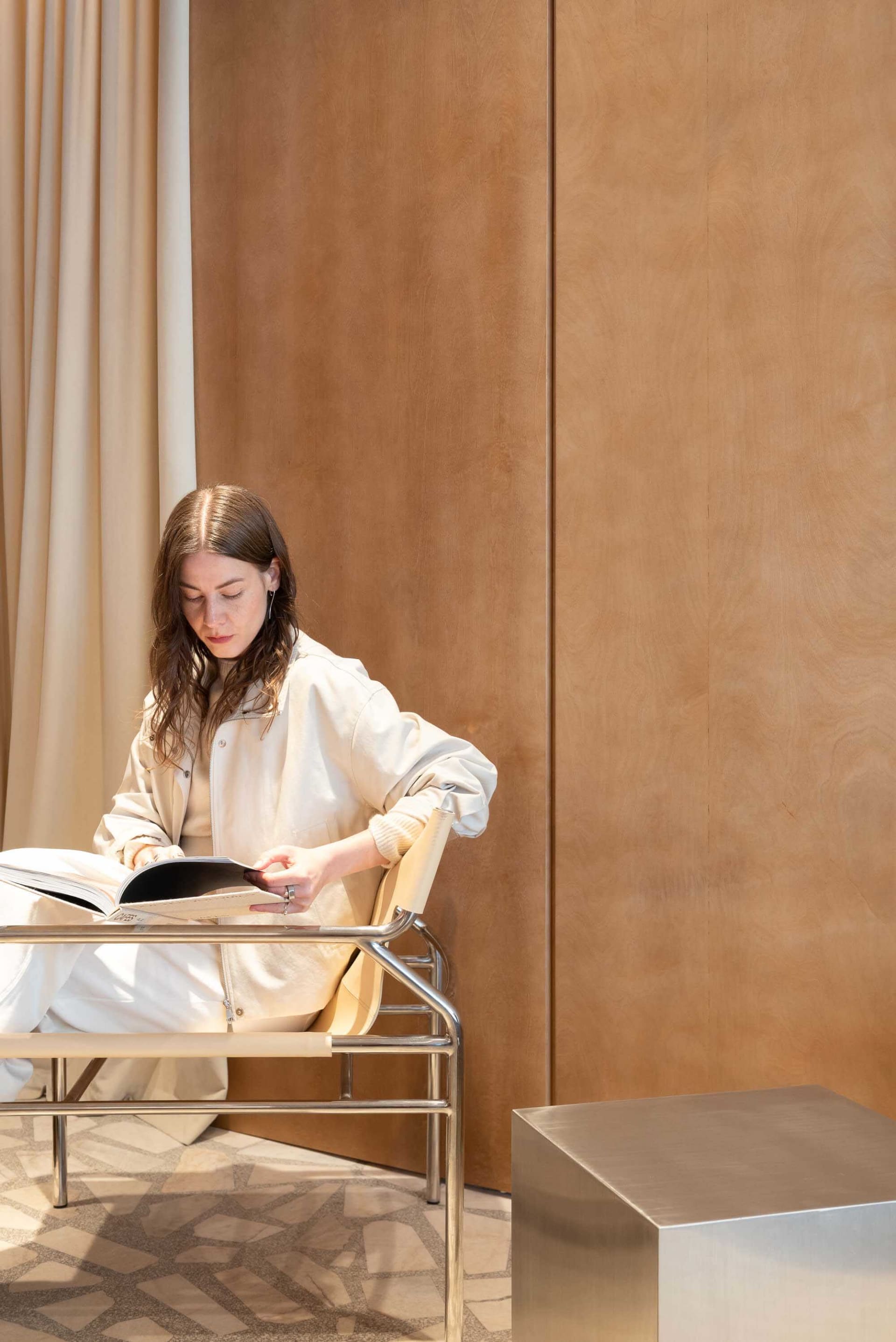 Woman with long brown hair reading a book while seated in a modern chrome and leather chair against a warm wood-paneled wall