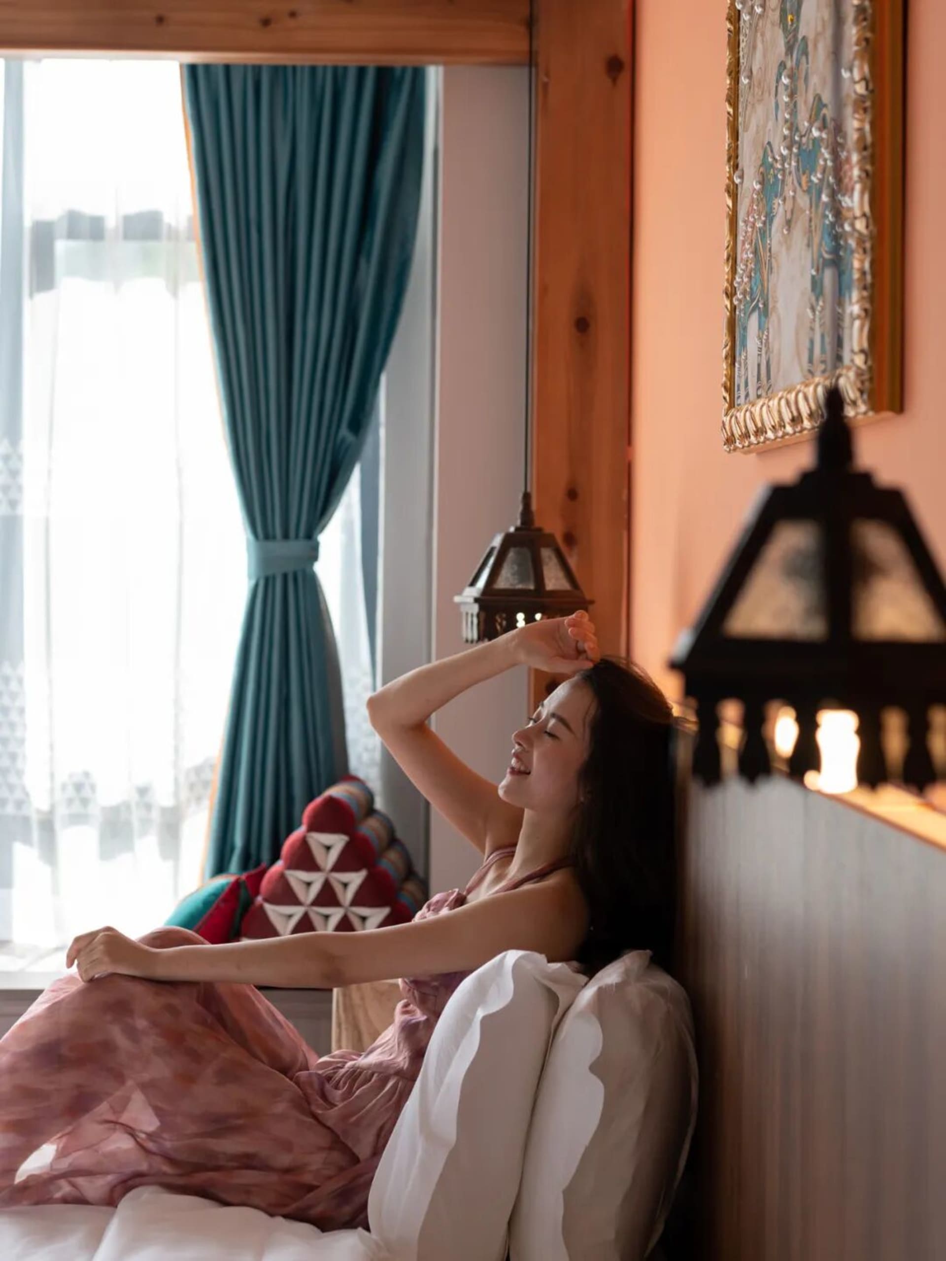 A young woman with dark hair in a pink dress reclines on a bed with white pillows, one hand behind her head, smiling with eyes closed in a cozy bedroom featuring wooden walls, teal curtains, and warm lantern lighting.