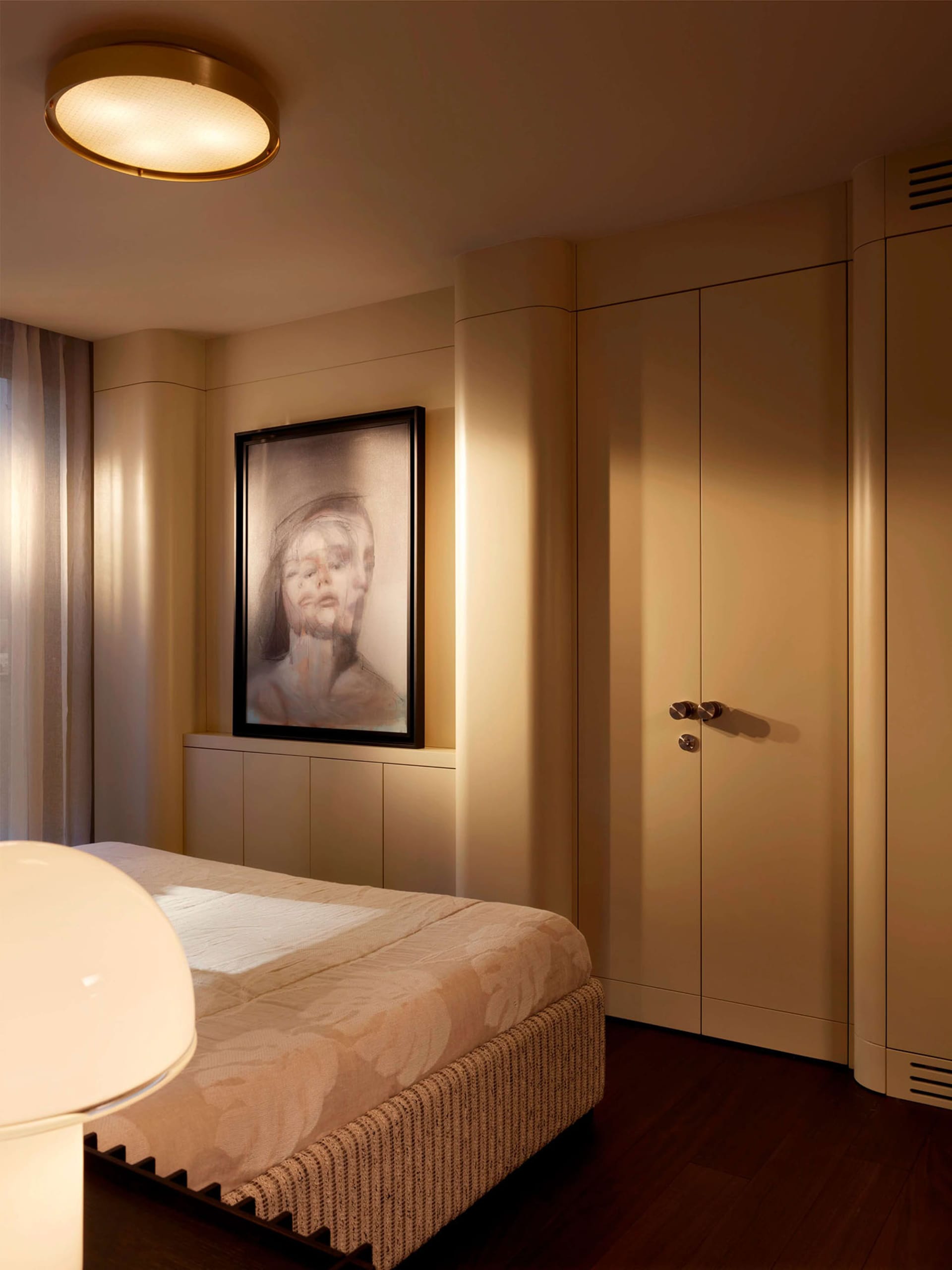 Modern minimalist bedroom featuring a built-in beige wardrobe, upholstered bed with floral coverlet, large portrait painting, and circular ceiling light fixture.