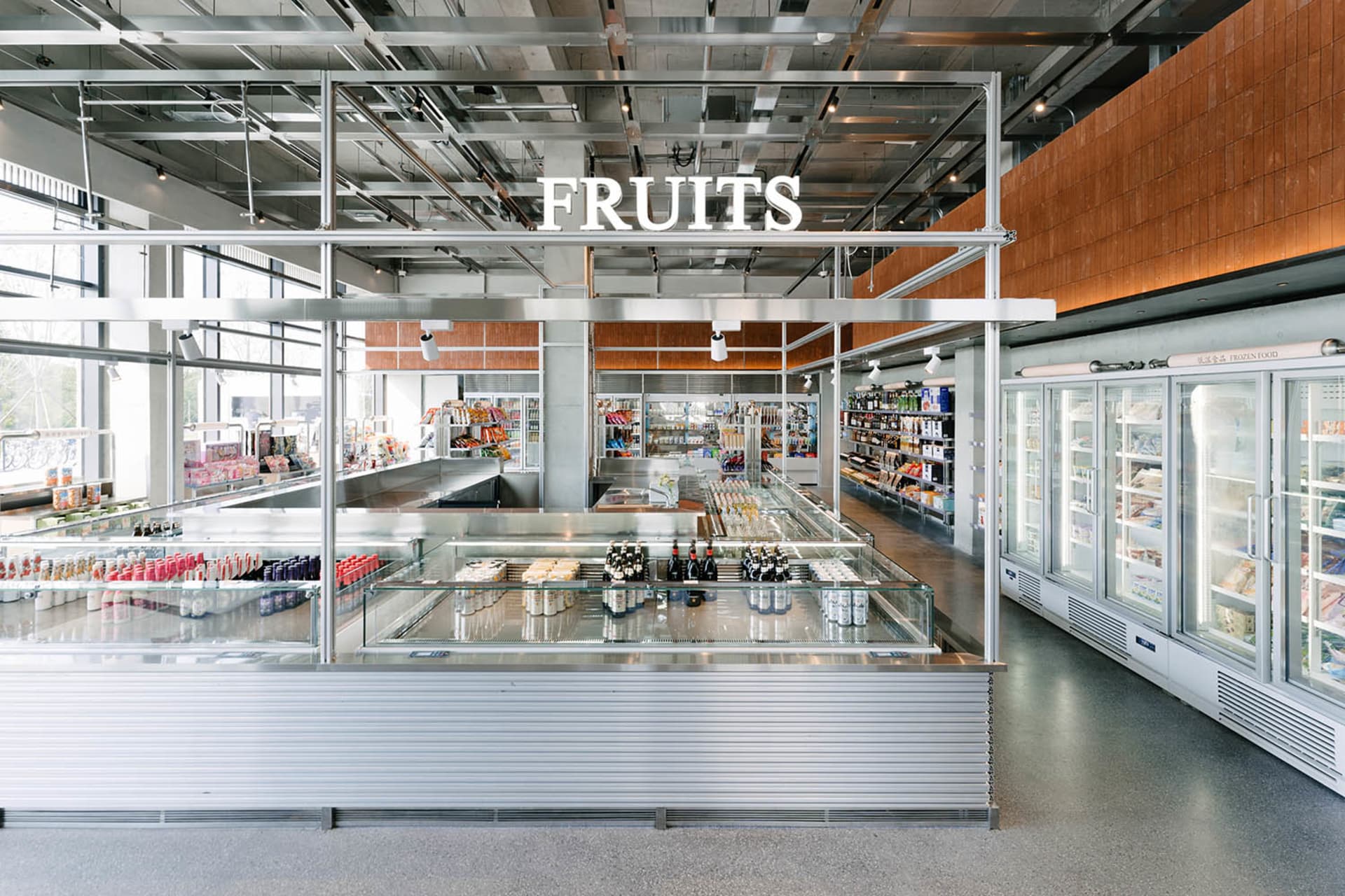 Wide view of a modern industrial-style grocery store featuring a large stainless steel central display island, glass refrigerated cases, concrete floors, and exposed ceiling infrastructure with a prominent FRUITS sign.