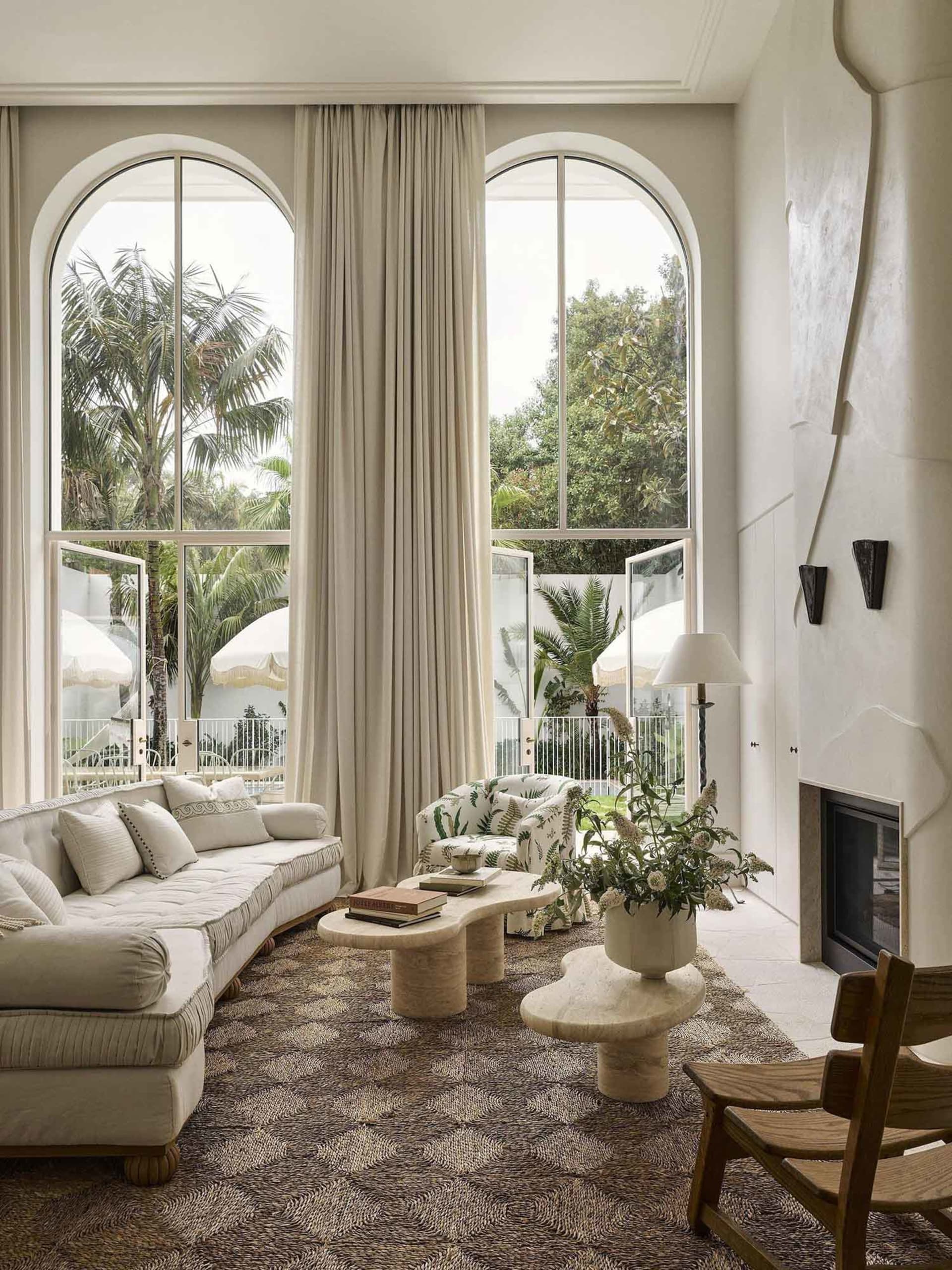 Bright living room featuring a curved white sofa, patterned rug, organic travertine coffee tables, a tropical print armchair, and large arched windows overlooking a palm-filled garden.