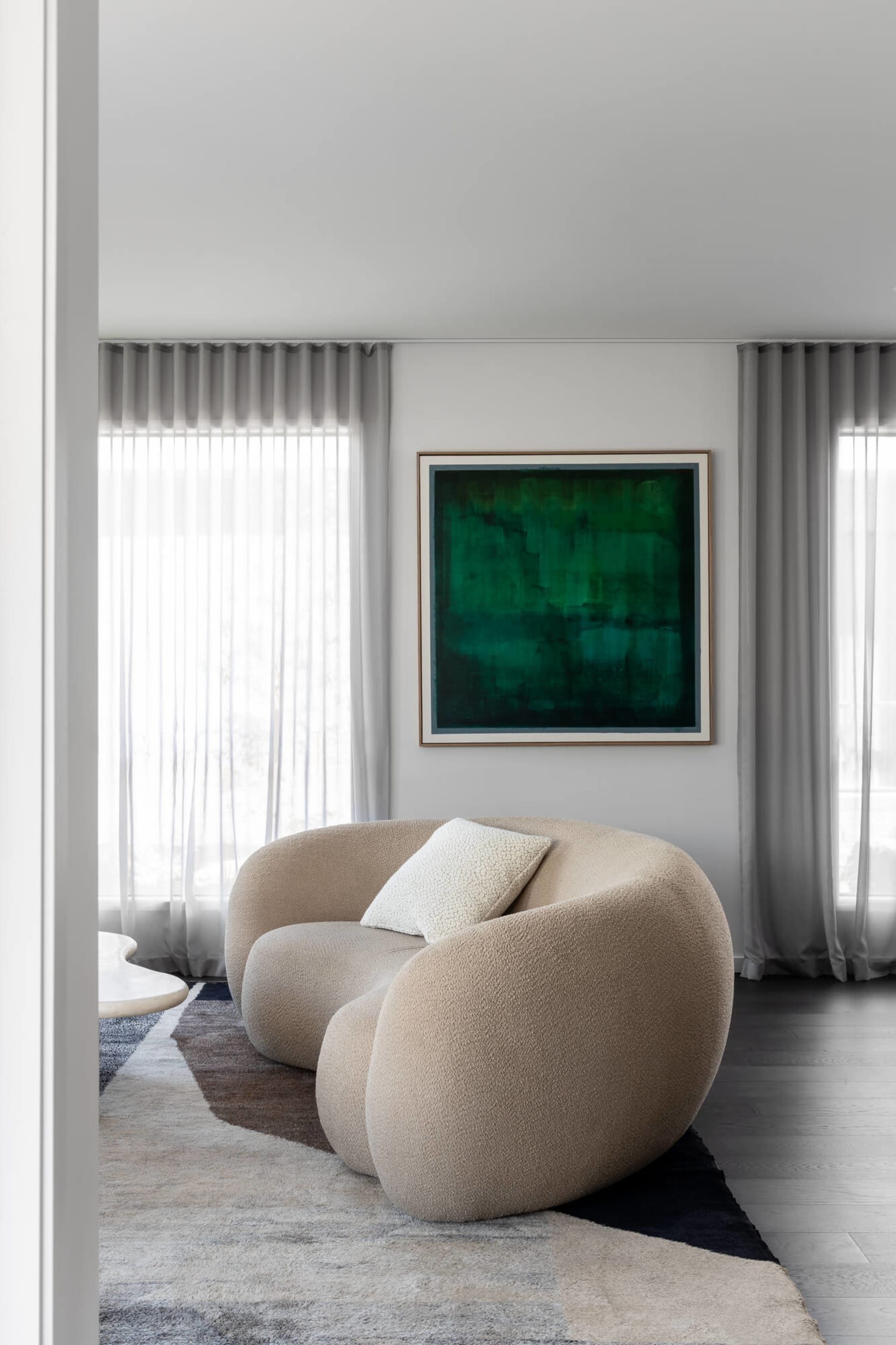 Modern living room featuring a curved beige boucle sofa with a white textured pillow, positioned on a geometric area rug beneath a large emerald green abstract painting, with sheer grey curtains in the background.