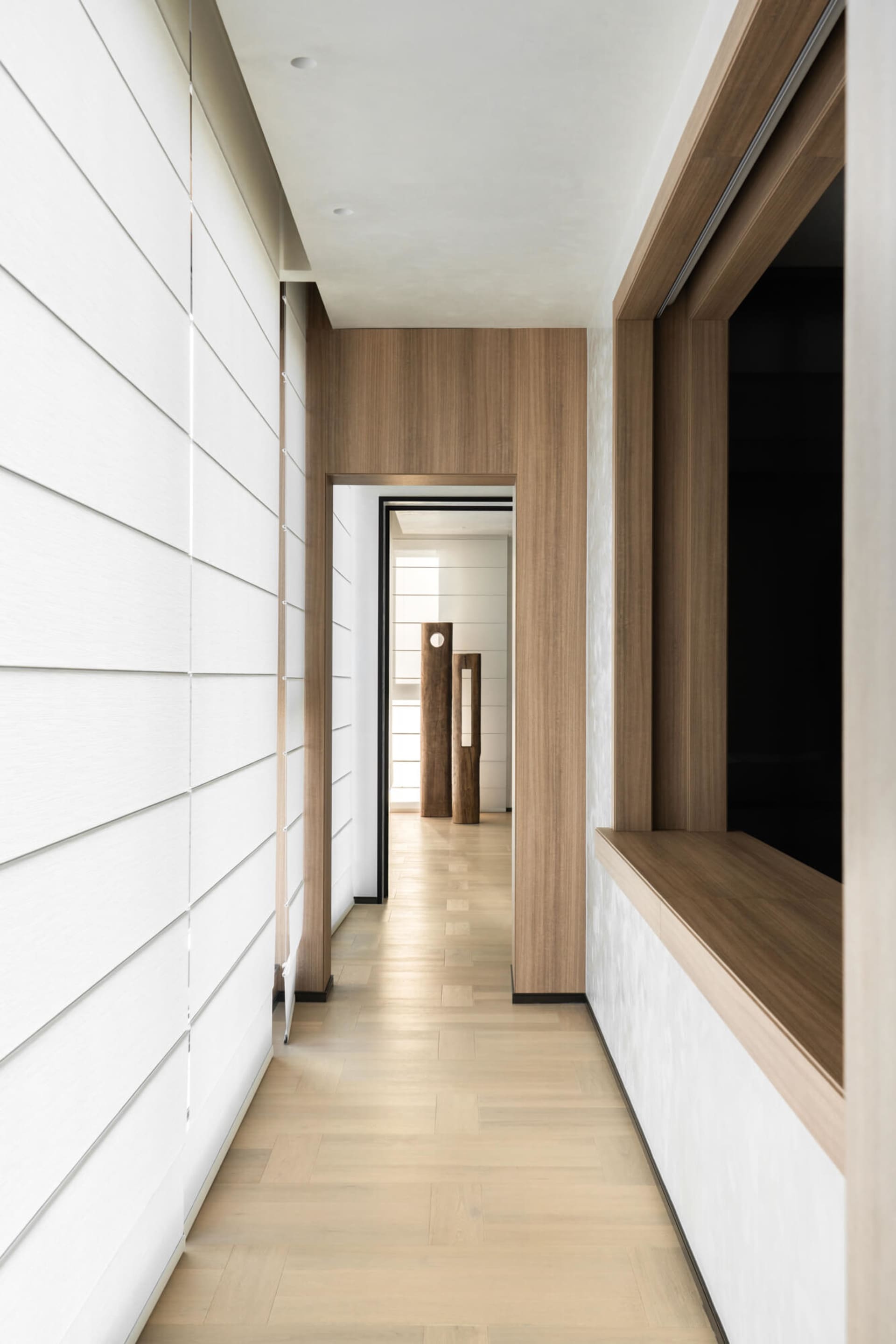 Modern minimalist hallway featuring white horizontal wall paneling, light oak wood accents, built-in window bench, and sculptural wooden pillars visible in the distance.