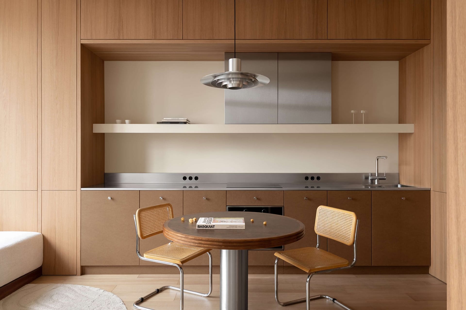 Modern minimalist kitchen and round dining table with two cane chairs, warm oak cabinetry, stainless steel countertop and range hood, floating shelf, and sculptural pendant light.