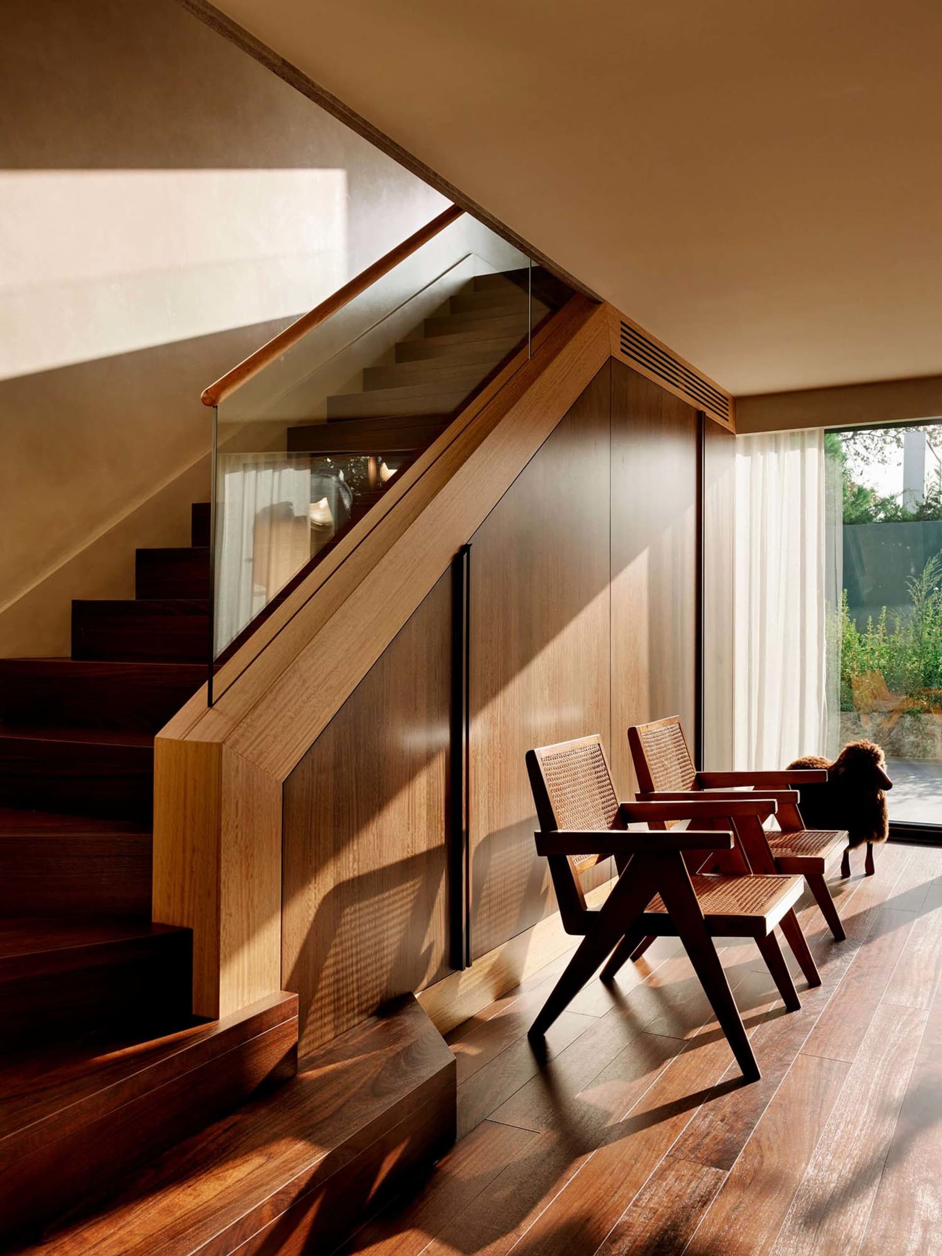 Modern interior featuring a dark walnut staircase with glass railing, built-in under-stair wooden storage, and three wooden rattan armchairs placed in a row on a wooden floor next to a large window.