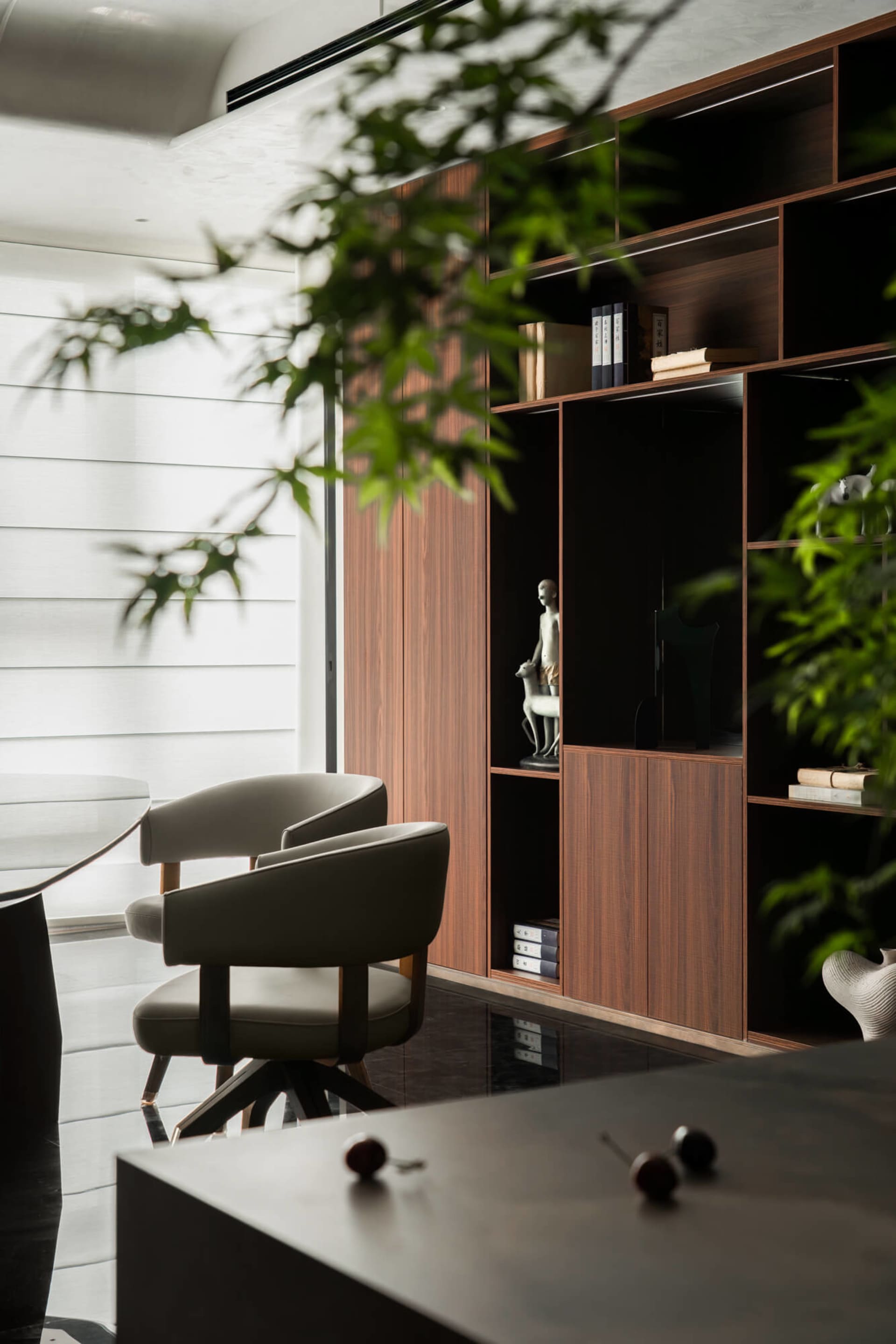 Modern dining area featuring two beige upholstered swivel chairs, a dark dining table, and a tall walnut built-in shelving unit displaying art objects, with lush green plants in the foreground.