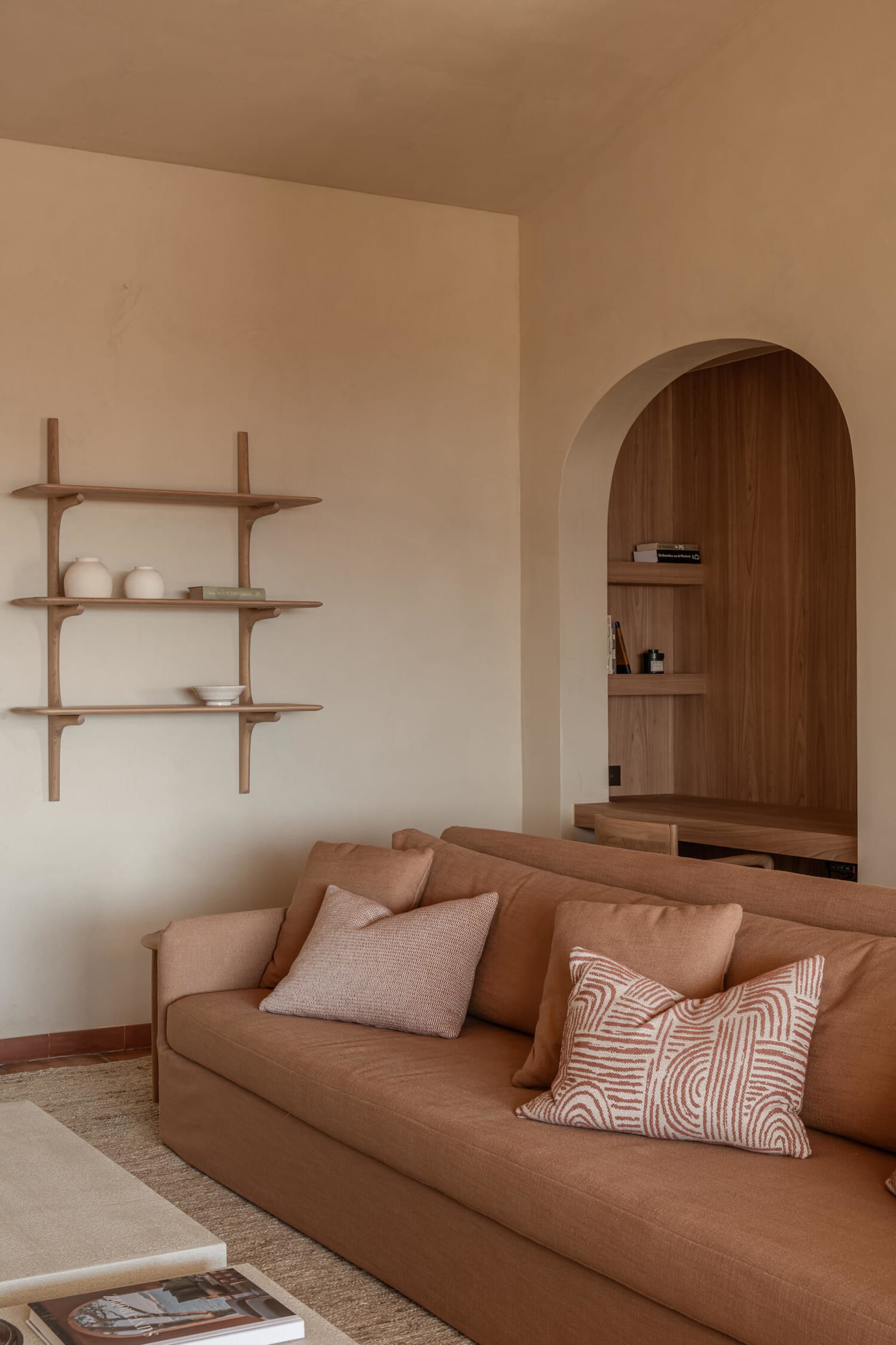 Modern minimalist living room with a terracotta fabric sofa, patterned cushions, floating wooden shelves, and a built-in arched wooden niche desk area.