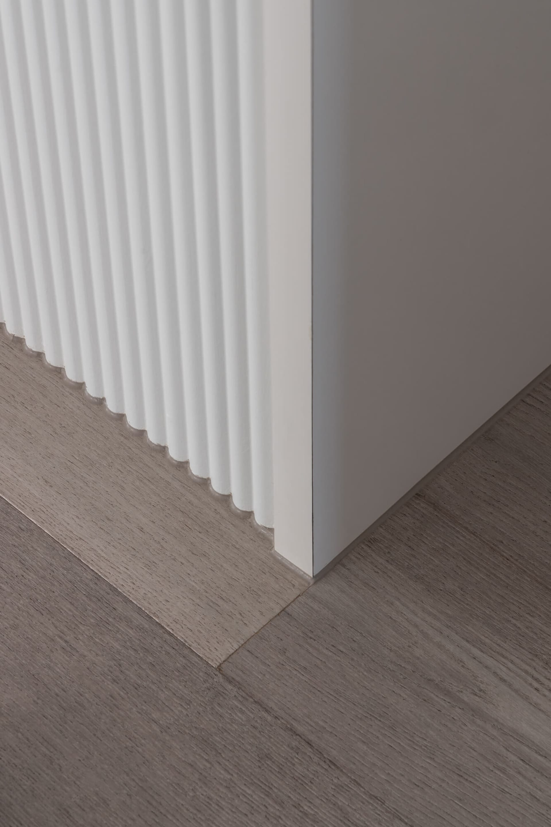 Close-up view of a white fluted baseboard meeting a grey-brown wood laminate floor at a clean 90-degree angle.