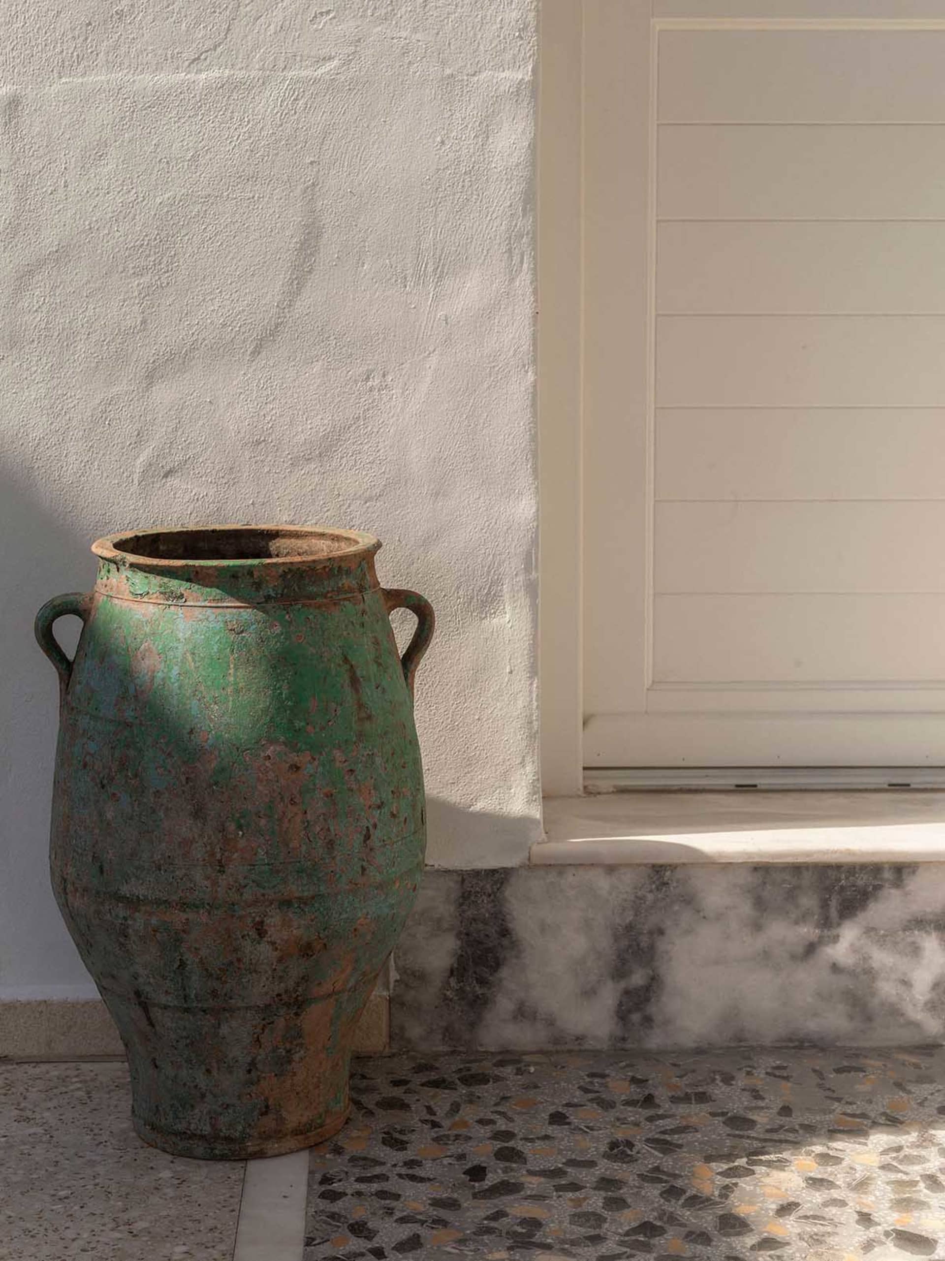 Large antique green ceramic urn with two handles placed on a terrazzo floor next to a white textured wall and marble base.