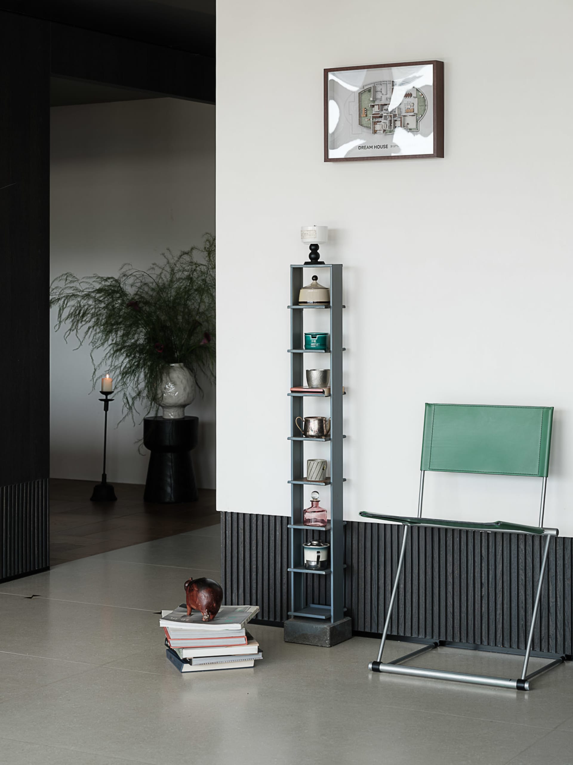 Modern minimalist entryway featuring a green leather folding chair, tall slate-blue shelving unit with ceramics, framed floor plan artwork, and a stack of books with a wooden hippo sculpture on a polished concrete floor.