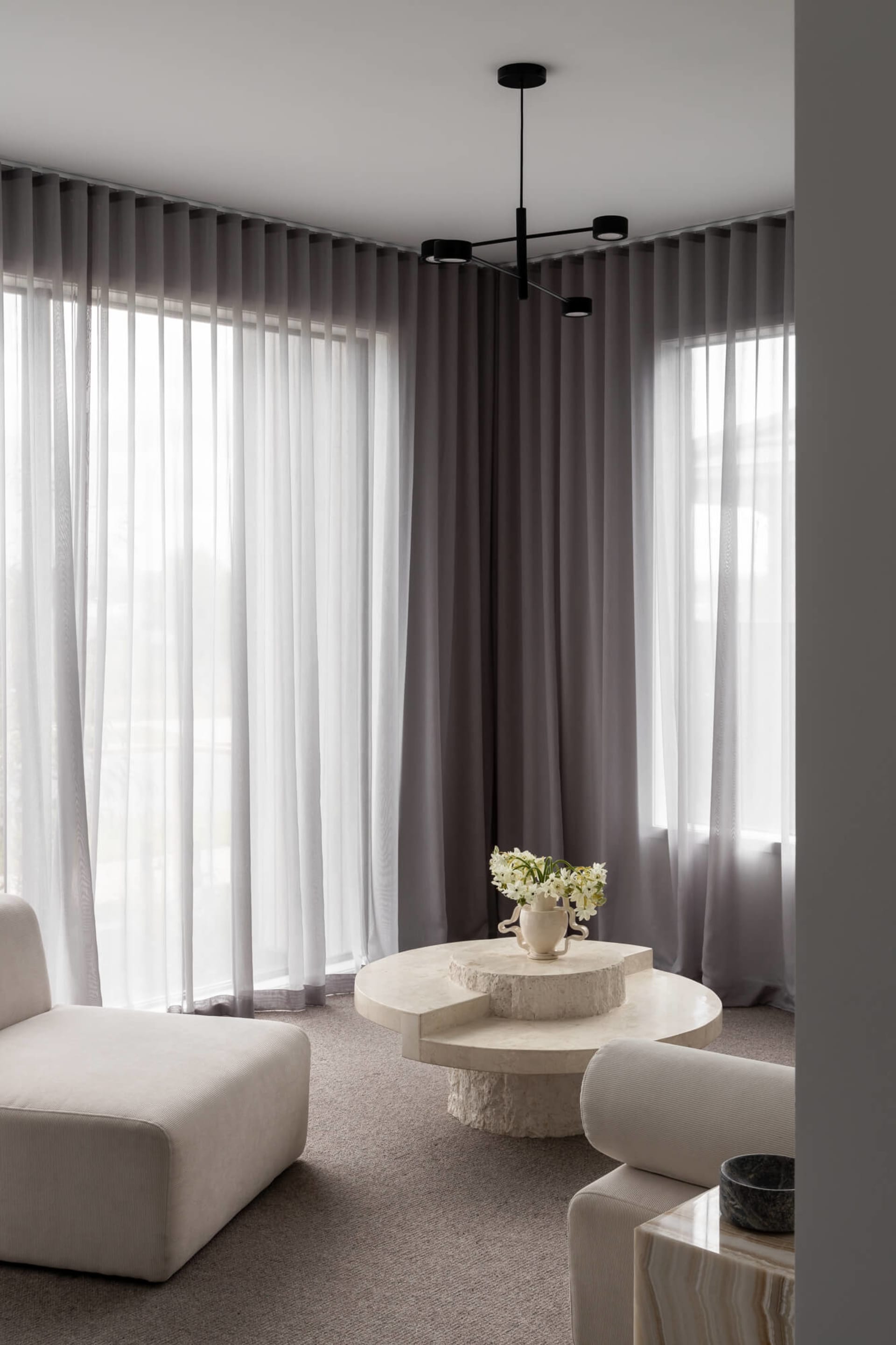 Minimalist living room featuring a large irregular travertine coffee table, modular cream bouclé sofas, layered sheer and opaque grey curtains, and a modern black ceiling light fixture.