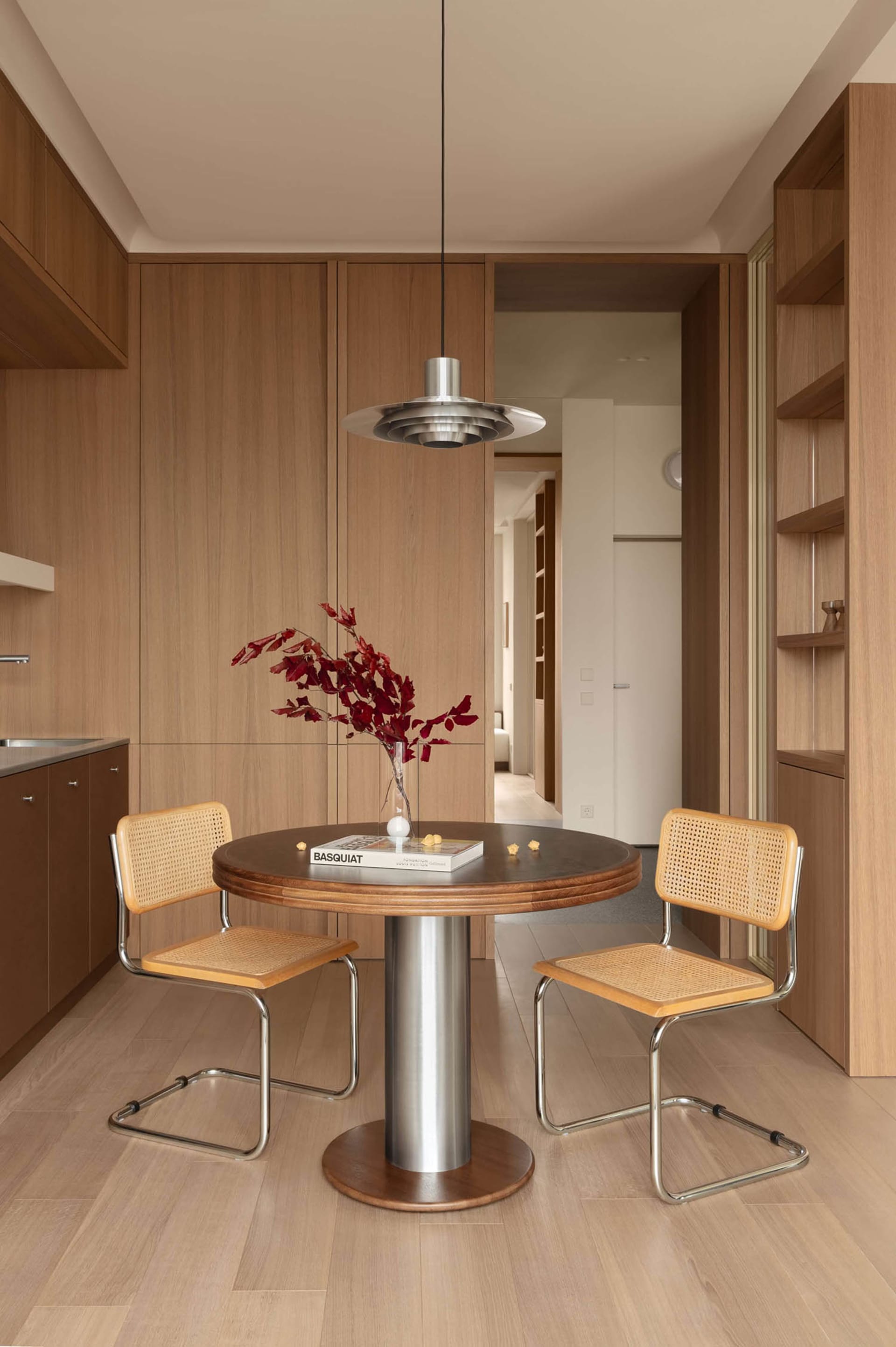 Modern minimalist dining area featuring a round walnut table, two cane chairs with chrome frames, a sculptural metallic pendant lamp, and a vase with red branches against warm wood cabinetry.
