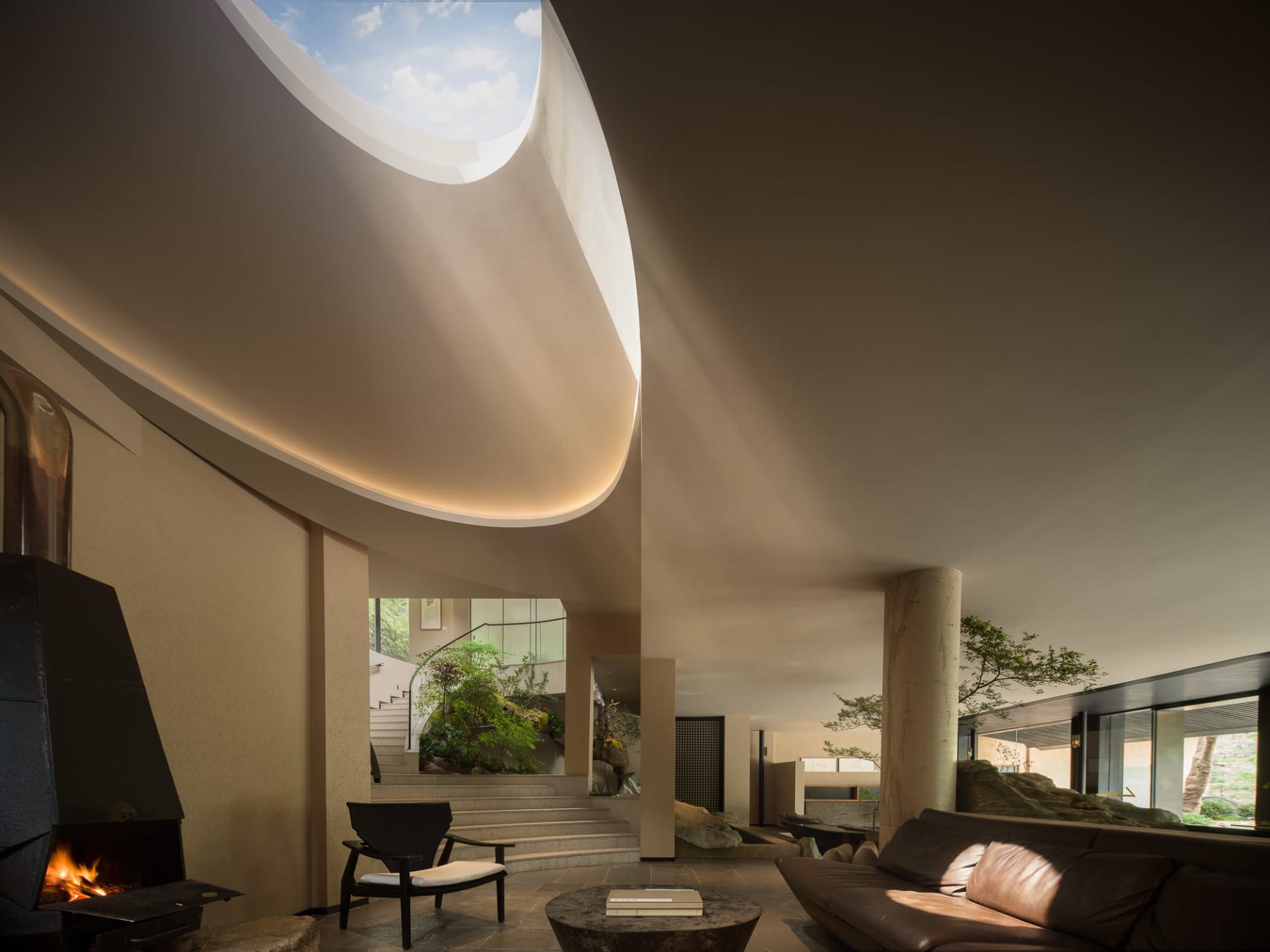 Modern minimalist living room featuring a massive curved skylight, concrete walls, a black fireplace, brown leather sofa, and an indoor courtyard with rocks and trees.
