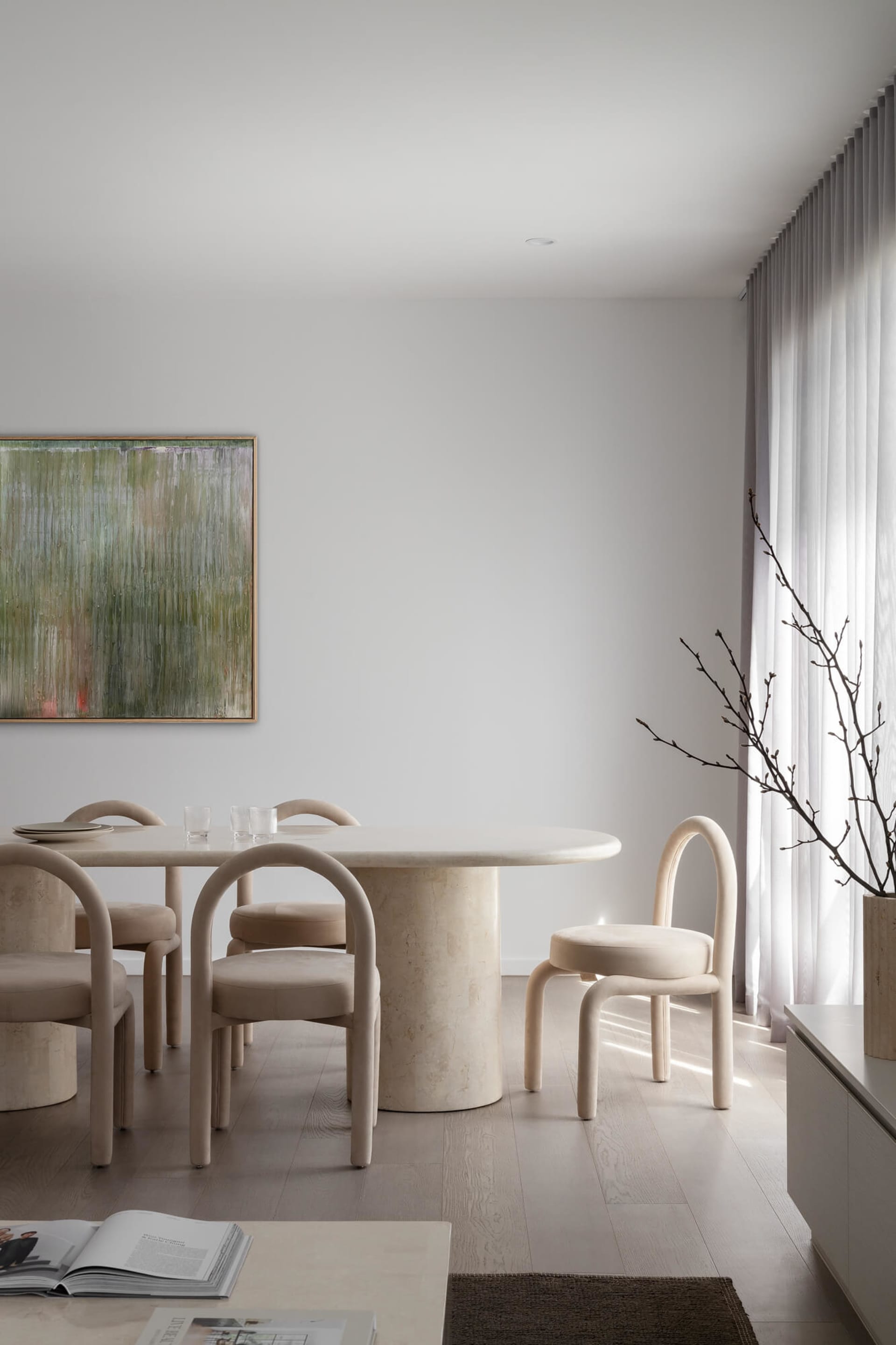 Minimalist dining room featuring a round travertine pedestal table, six curved beige upholstered chairs, large abstract green painting, and sheer curtains allowing natural light.