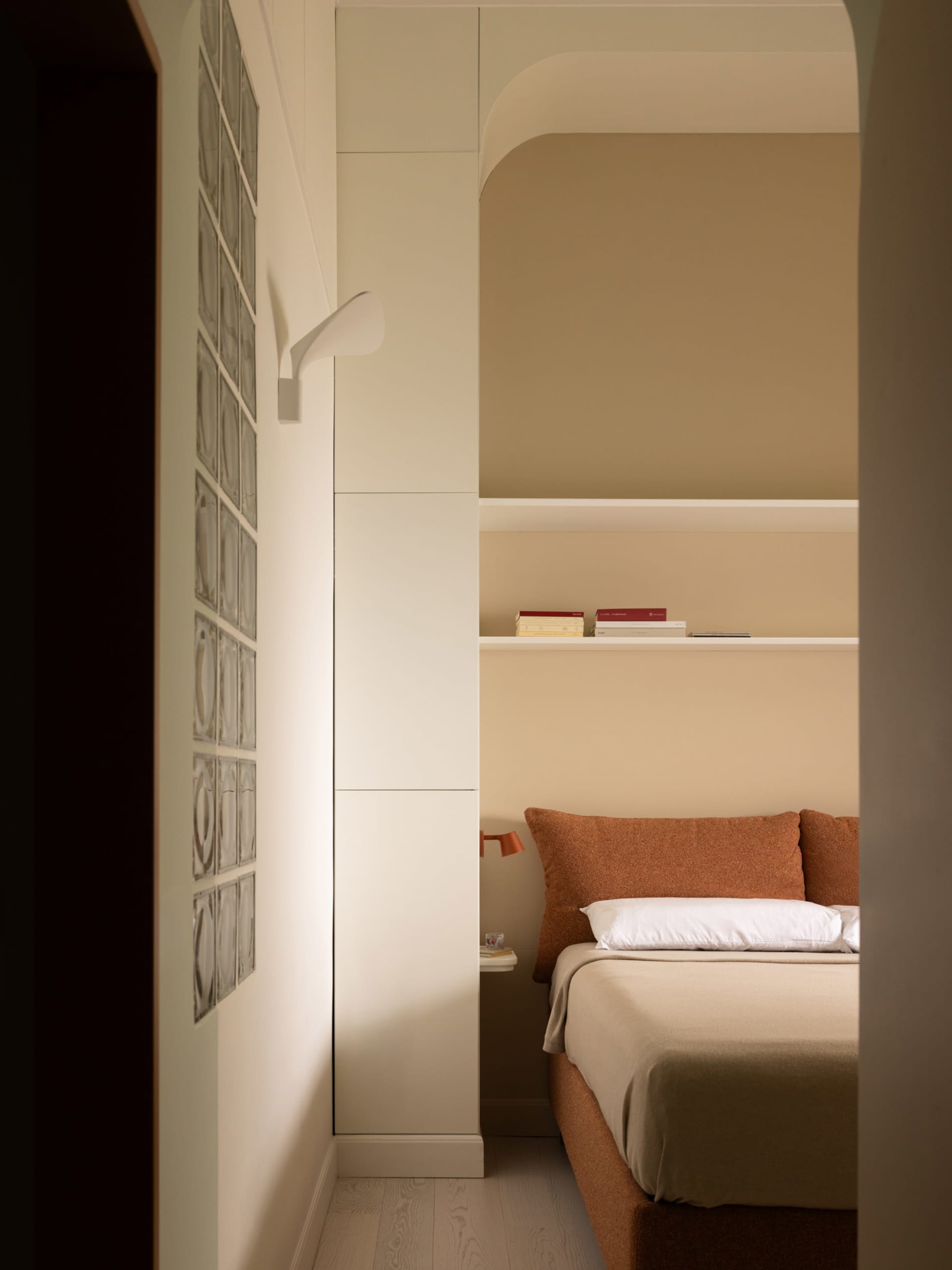 View into a compact modern bedroom featuring a terracotta boucle bed with white linens, built-in arched shelving, and a glass block wall under warm ambient lighting.