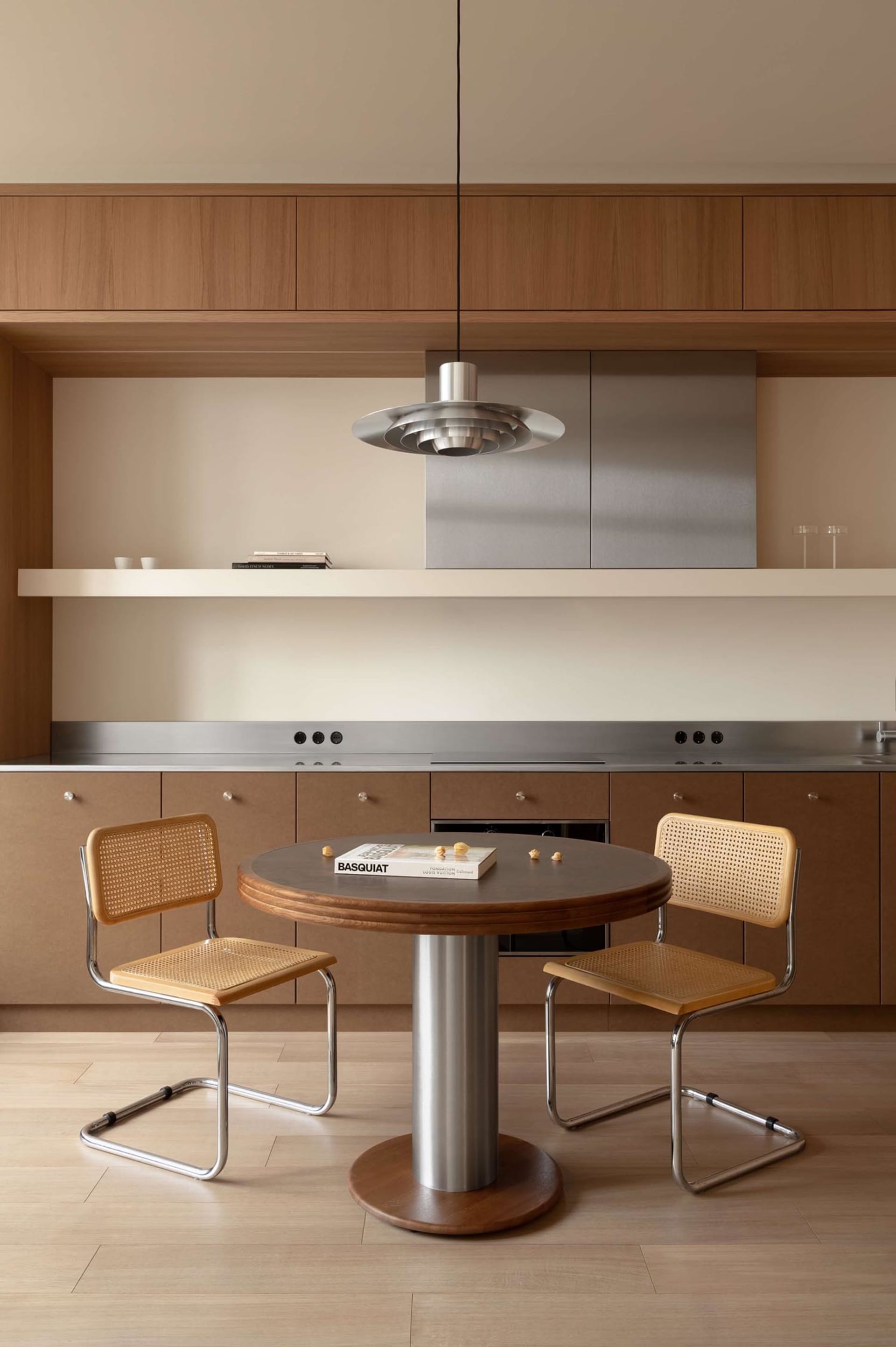 Modern minimalist dining area with a round walnut table, two cane and chrome chairs, stainless steel kitchen counter, and wooden upper cabinets under a sculptural metallic pendant lamp.