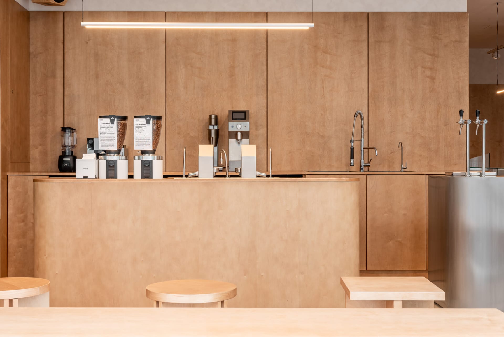 Modern minimalist coffee bar made of light birch plywood with built-in espresso machines, grinders, and stainless steel taps, illuminated by a long linear pendant light.