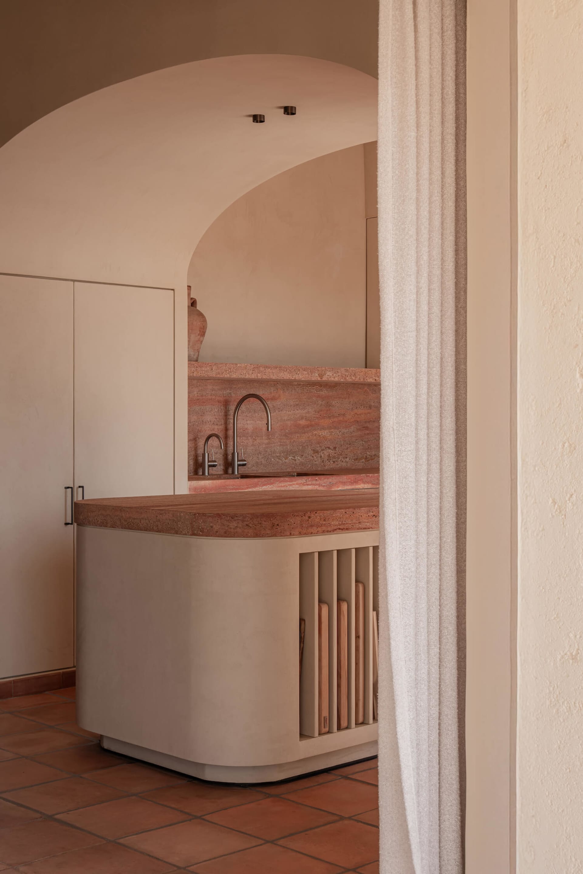 View through a linen curtain into a modern kitchen featuring a curved island with terracotta countertop, plaster base, and wooden slat storage, set against a large arched opening and terracotta floor tiles.
