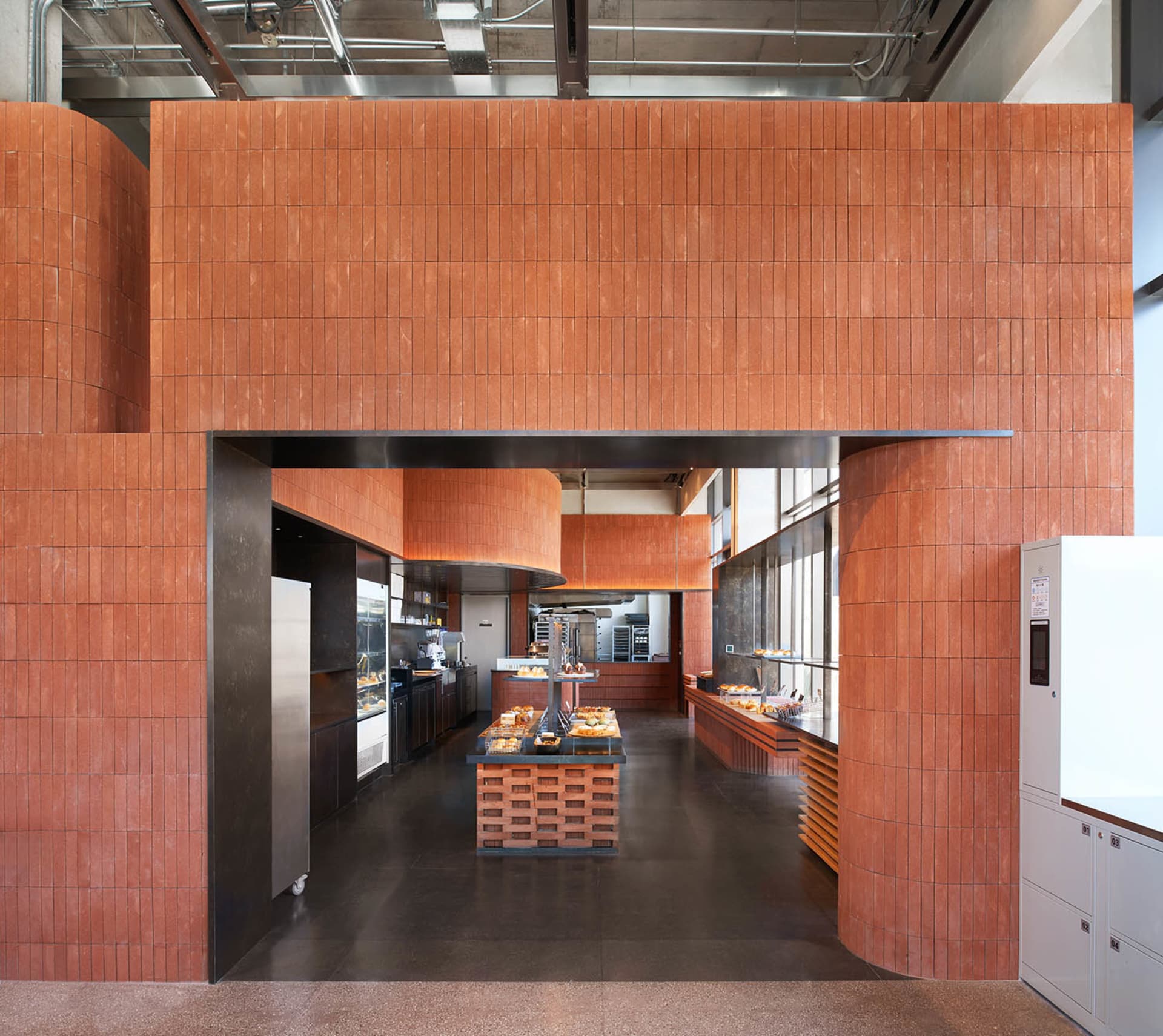 Modern bakery interior featuring extensive terracotta tile walls, a central wooden display island, dark concrete floor, and industrial ceiling with exposed ductwork.