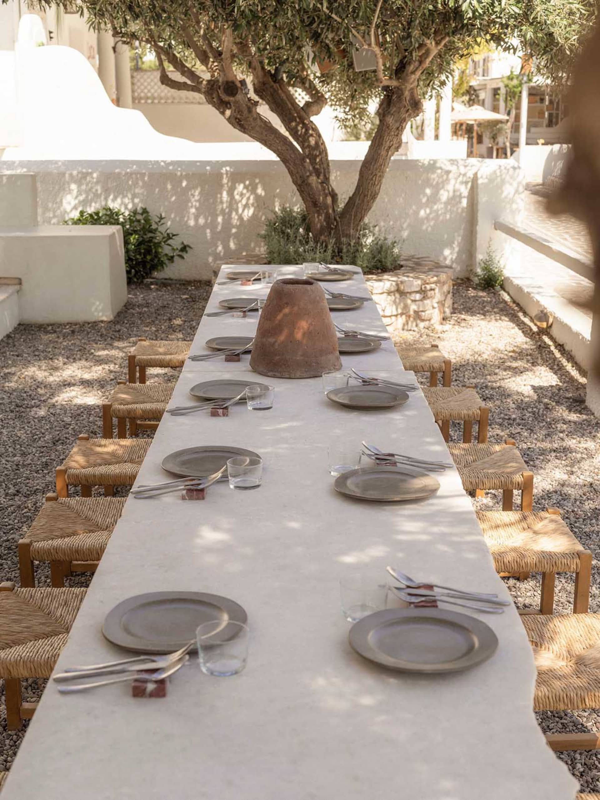Long outdoor dining table made of light stone with woven rush stools, set with ceramic plates and a large terracotta pot centerpiece in a gravel courtyard under an olive tree.