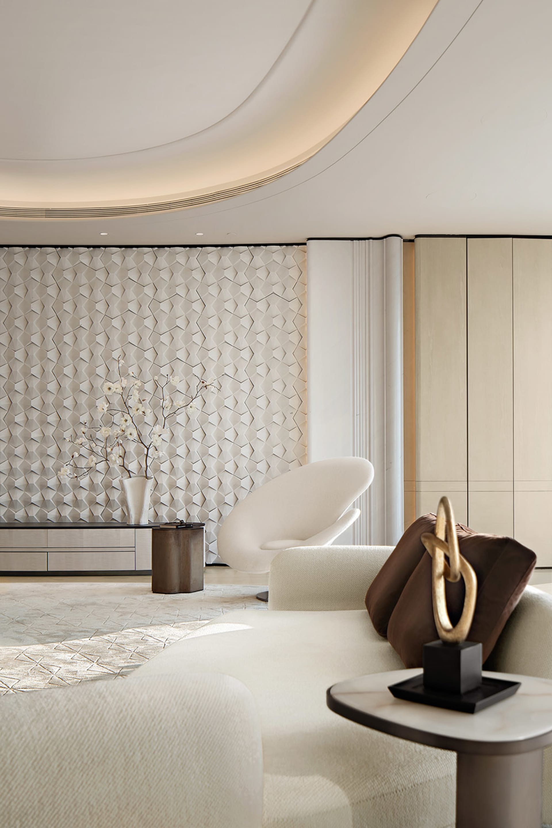 Modern luxury living room featuring a dramatic white 3D textured geometric wall, curved white lounge chair, cream sectional sofa with brown velvet pillows, and a sculptural gold knot ornament.
