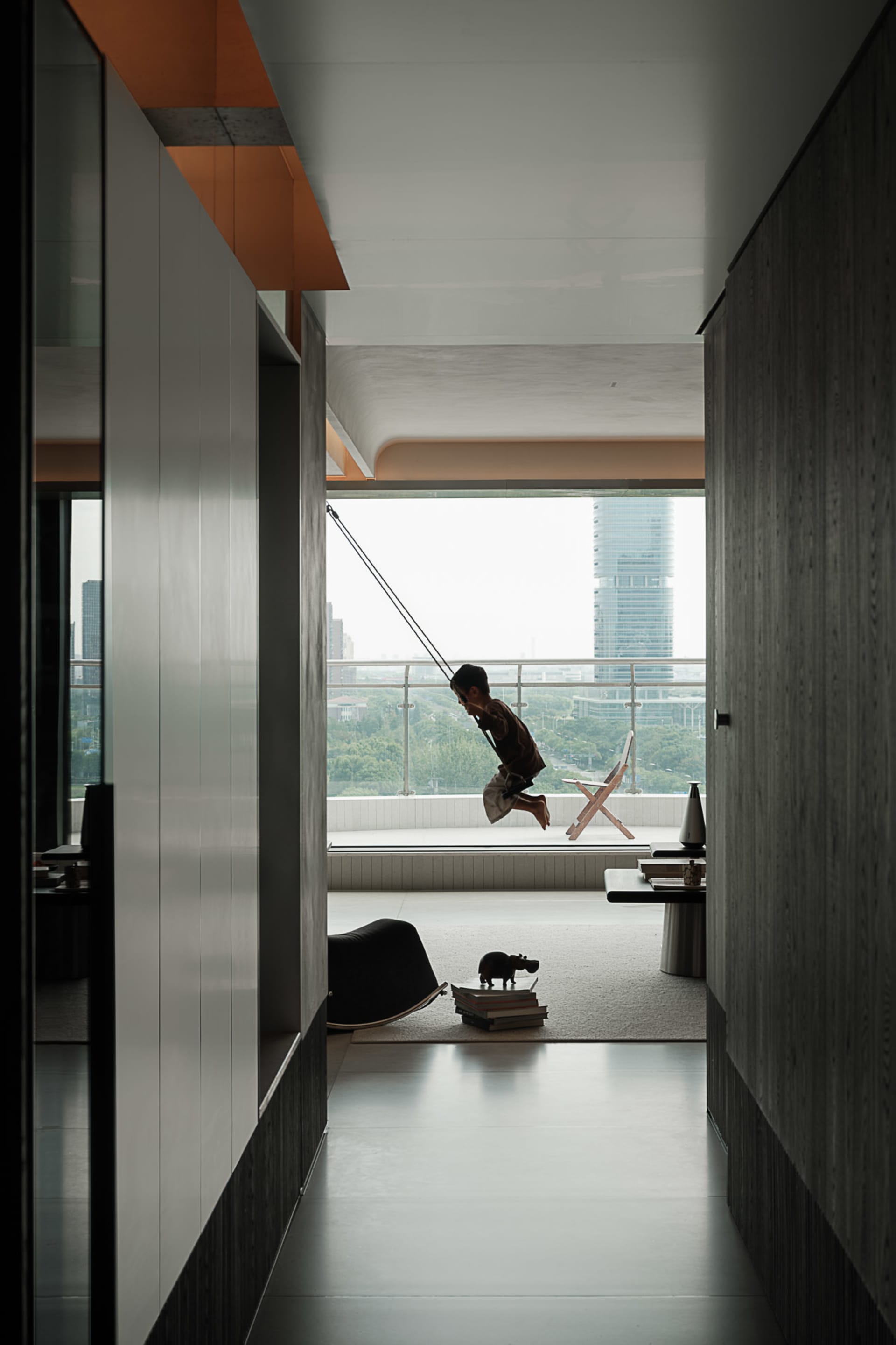 Modern minimalist apartment interior featuring a child on an indoor swing, concrete walls, large glass balcony overlooking a city skyline, and scattered children's toys on a light rug.