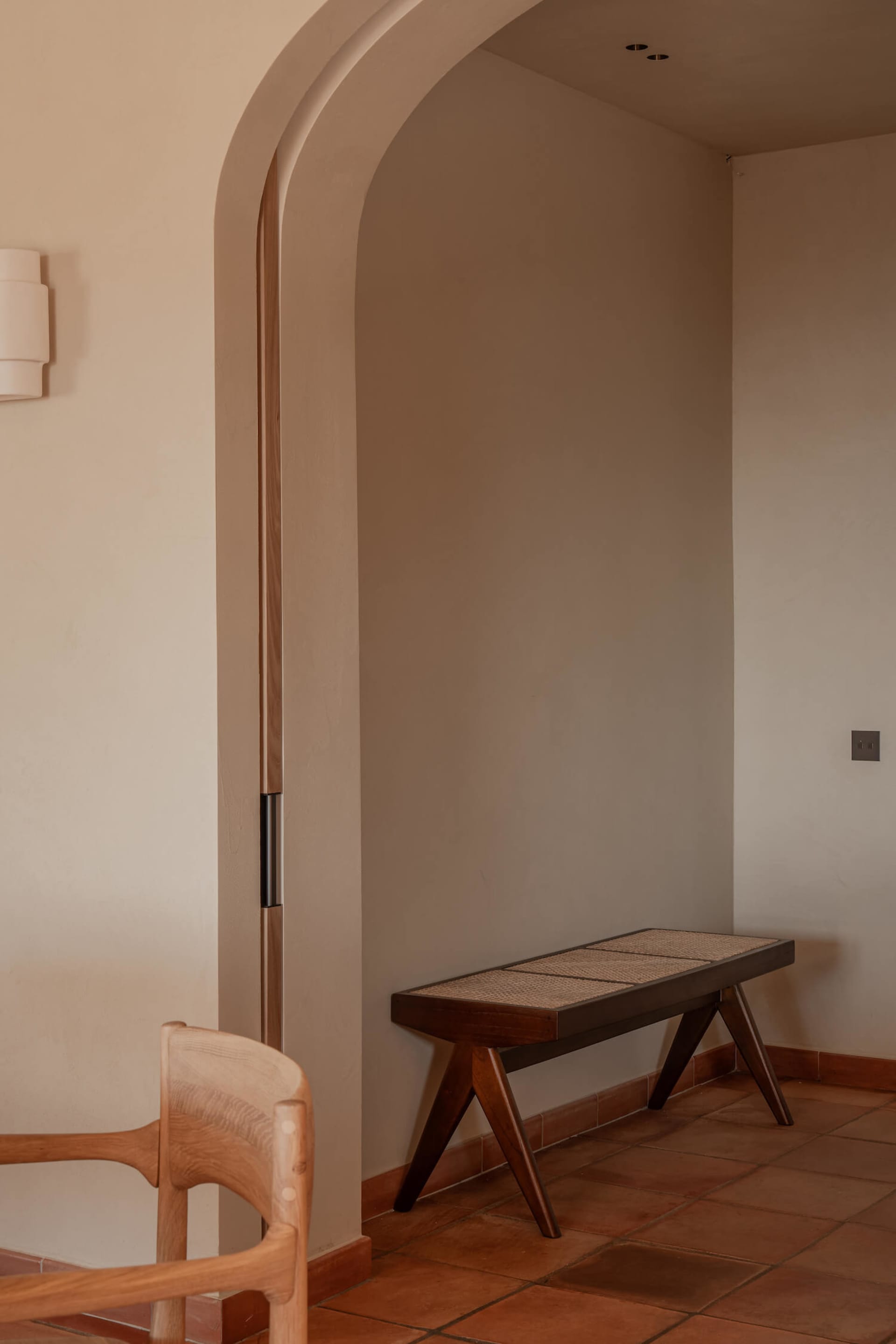 A serene entryway featuring a dark walnut bench with woven cane seat, terracotta floor tiles, arched doorway with wood trim, and a wooden chair in the foreground.