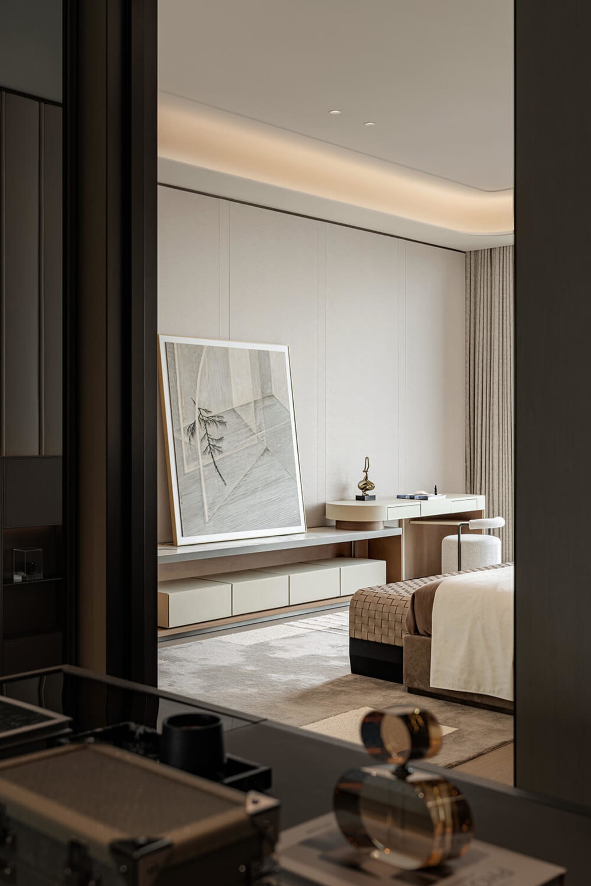 View into a minimalist luxury bedroom with neutral tones, a woven ottoman bench at the foot of the bed, a floating vanity desk with upholstered stool, and a large framed abstract line drawing on the wall.