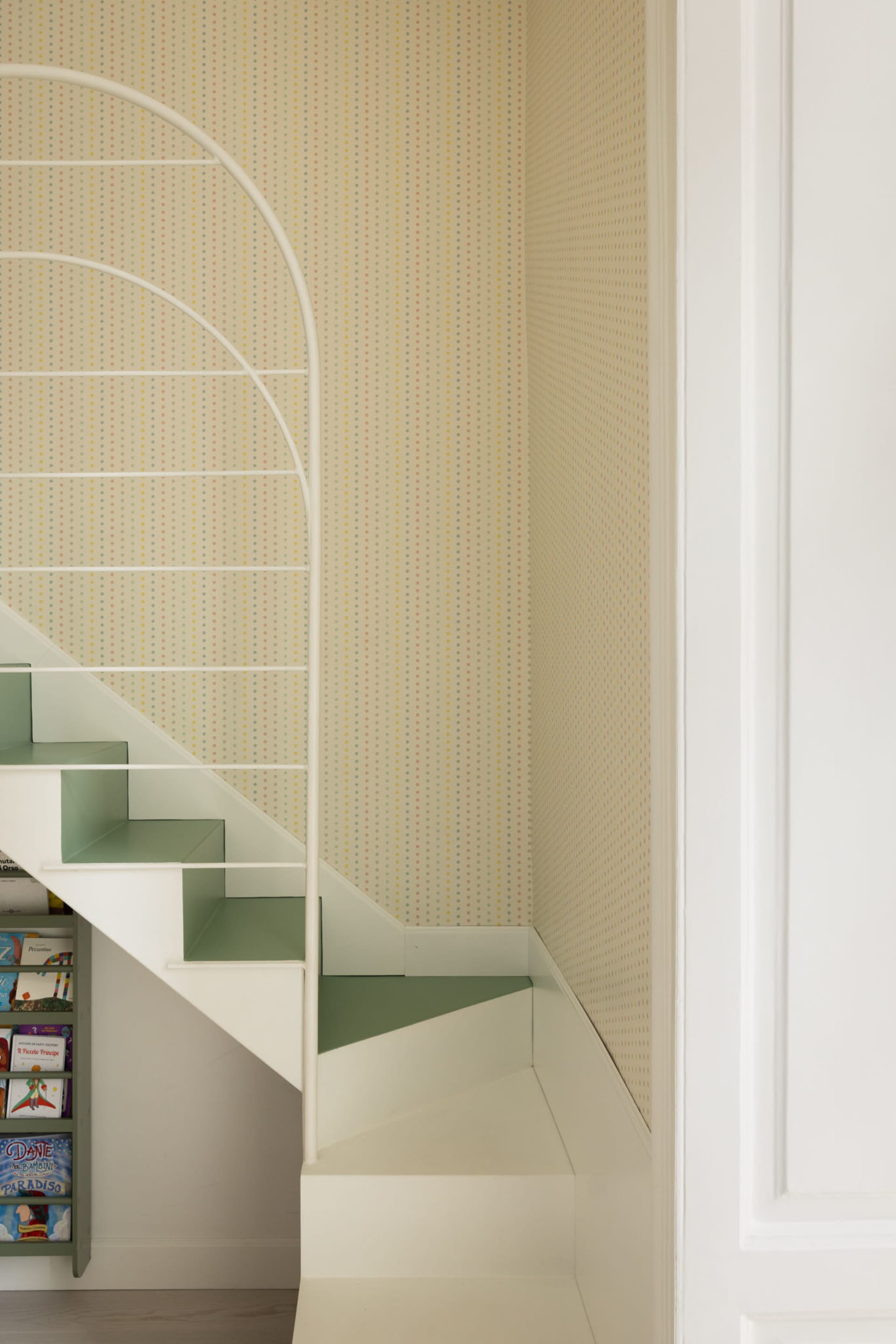 View of a modern children's staircase with sage green steps, white railing, and colorful polka dot wallpaper on the wall, with built-in bookshelf underneath.
